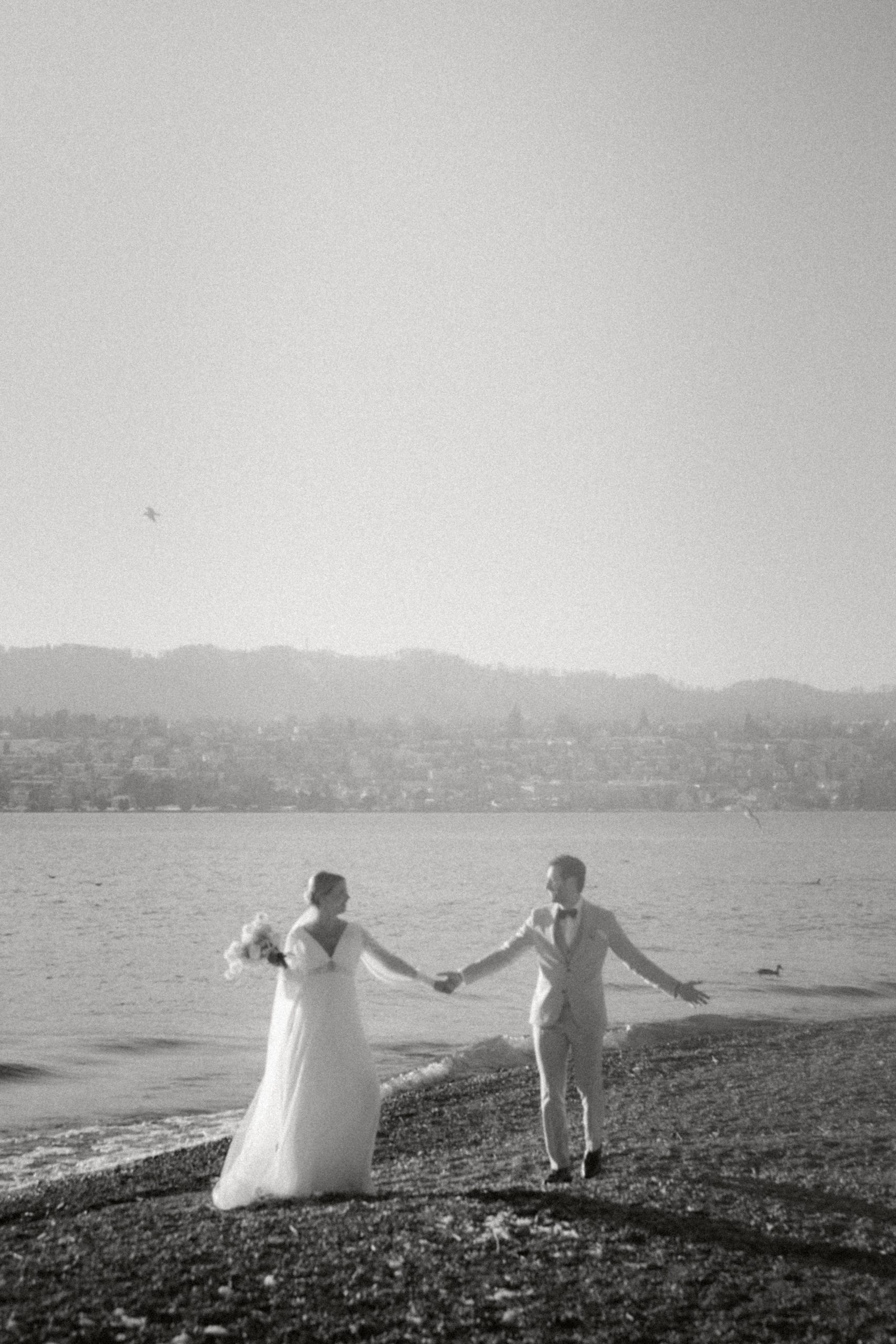 Braut und Bräutigam halten sich an den Händen und spazieren an einem Kiesstrand in der Nähe des Hotels See entlang, wobei sich das ruhige Wasser in der Sonne spiegelt. Das Schwarz-Weiß-Bild schafft eine zeitlose, romantische Hochzeitsatmosphäre mit fernen Hügeln im Hintergrund.