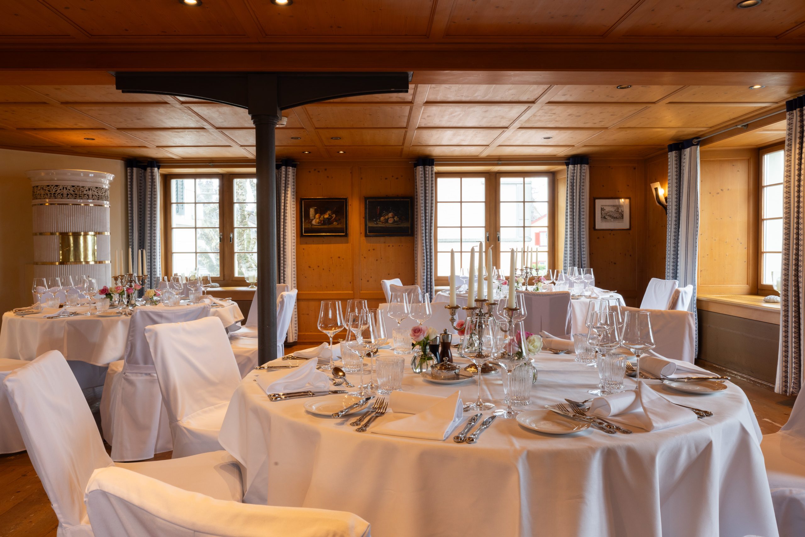 Eleganter Turmsaal im Event Hotel Sonne mit runden Tischen mit weißen Tischtüchern, Gläsern und Blumen. Warme Holzdecken und -wände, große Fenster mit Vorhängen und gerahmte Kunstwerke schaffen ein einladendes Ambiente.