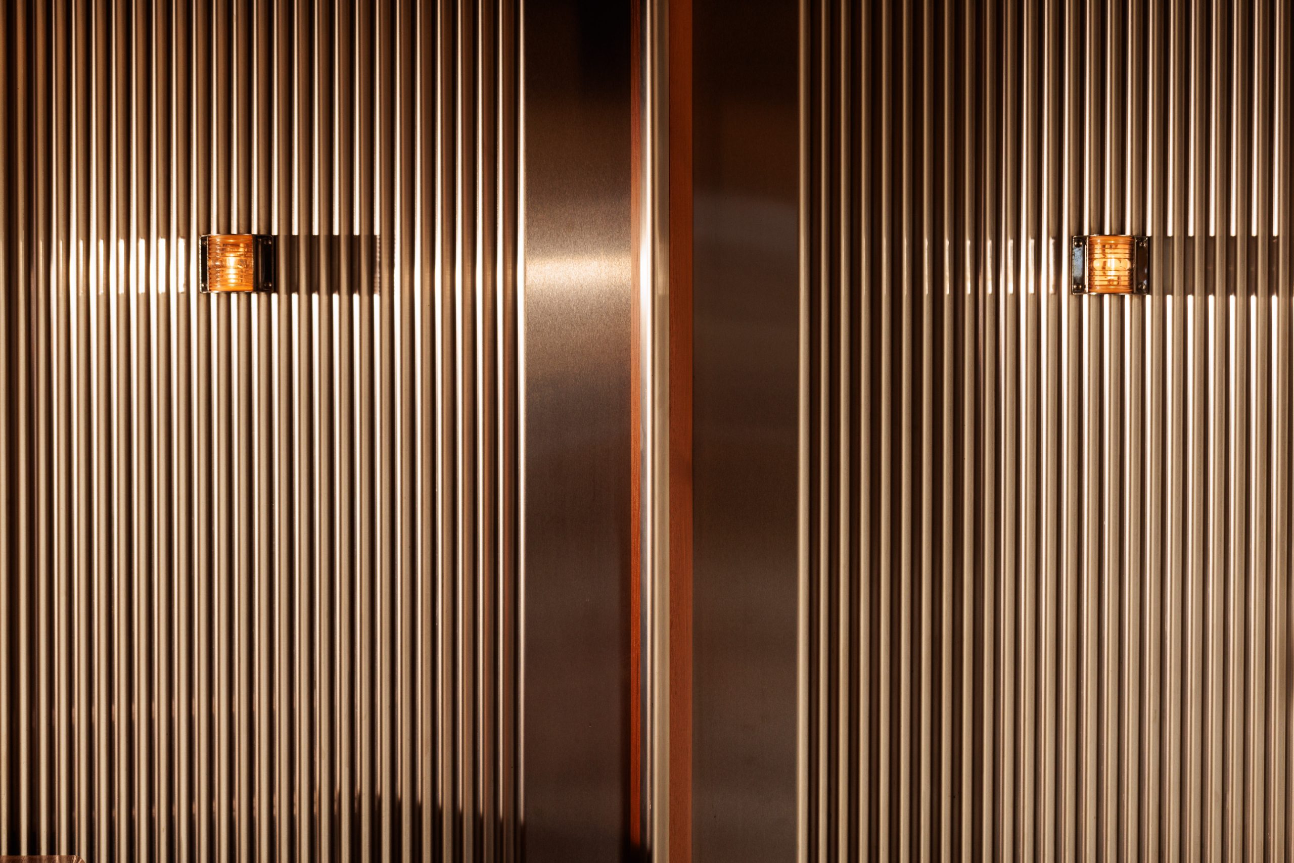 Nahaufnahme von zwei bronzefarbenen, vertikal gerippten Sonnengallerie-Metalltüren mit kleinen rechteckigen Lichtern in der Mitte, die markante Schatten auf die strukturierte Oberfläche werfen.