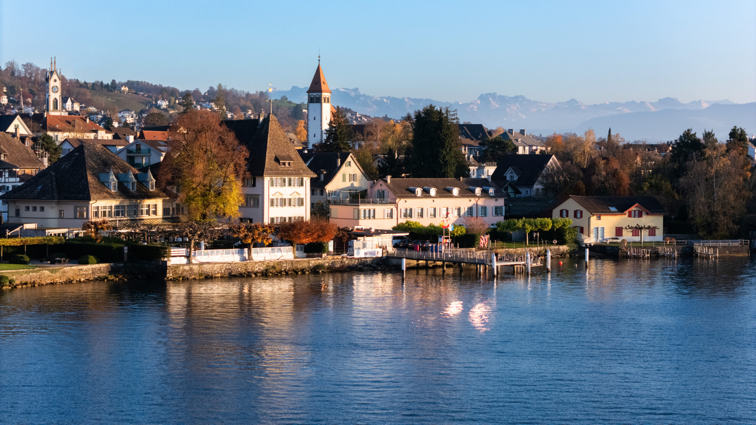 Ein malerisches Seedorf mit charmanten Häusern, dem Romantikhotel Sonne Küsnacht, einem Kirchturm, Herbstbäumen und schneebedeckten Bergen unter blauem Himmel, die sich im ruhigen Wasser des Sees spiegeln.