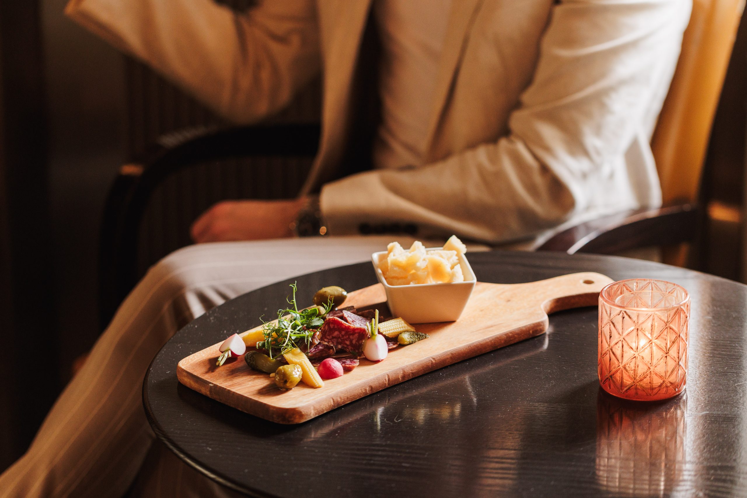 Eine Person in einem hellen Anzug sitzt an einem dunklen Tisch im Tapas Hotel Sonne Küsnacht, mit einem Holzbrett mit Gurken und Käse, einem kleinen Käseteller und einem orangefarbenen, strukturierten Glas.
