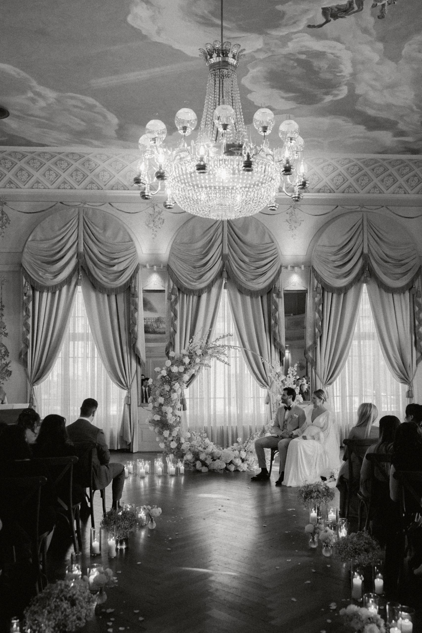 Ein Schwarz-Weiß-Foto von einer Hochzeit am Zürichsee in einem eleganten Raum mit einem Kronleuchter, großen drapierten Fenstern, einem Blumenbogen und Kerzen am Gang. Die Braut und der Bräutigam sitzen zusammen an der Stirnseite.