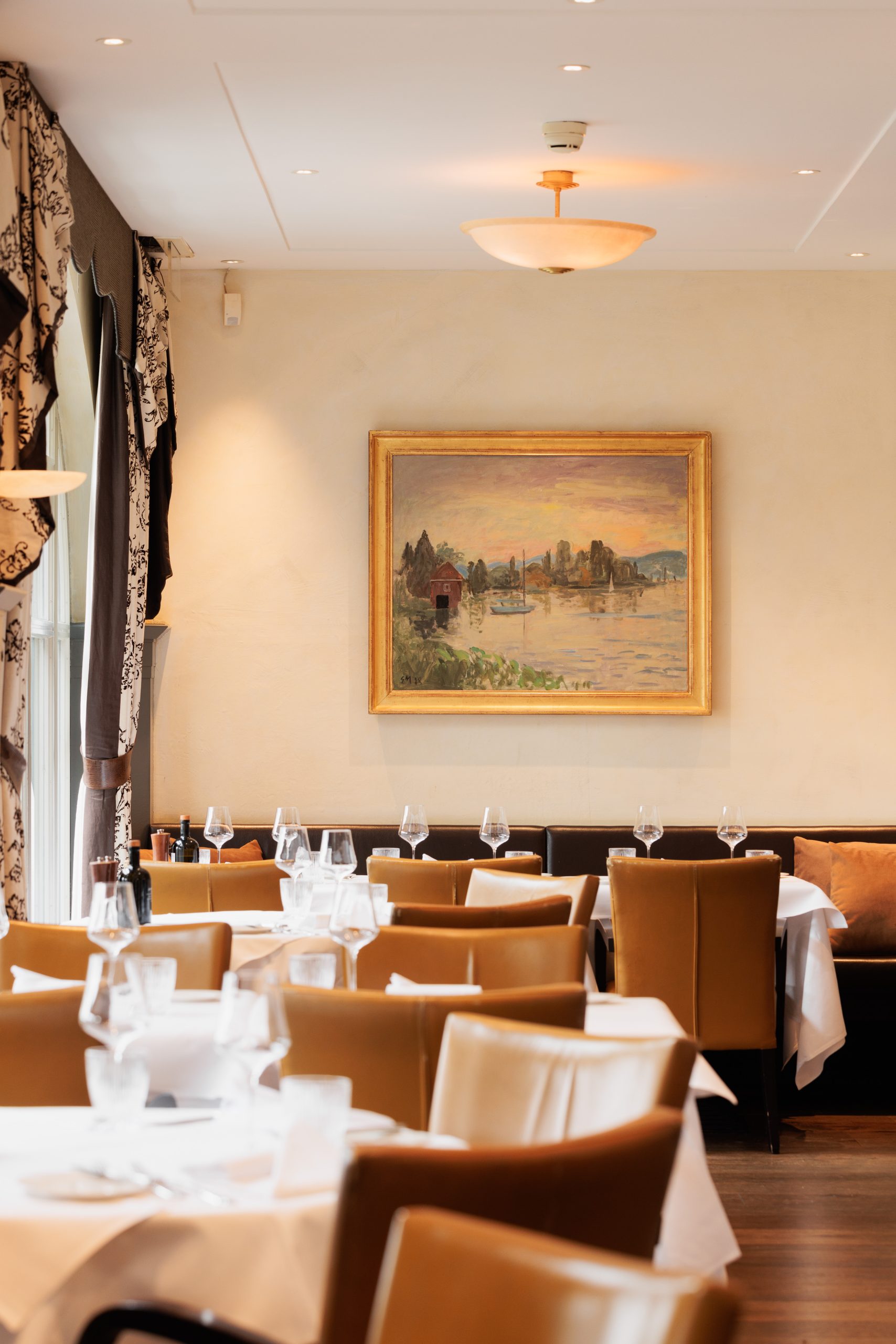 Ein heller Essbereich im Restaurant Sonnengalerie mit hellbraunen Lederstühlen, weißen Tischtüchern und ordentlich gedeckten Tischen. An einer cremefarbenen Wand über den Sitzplätzen hängt ein Landschaftsgemälde in einem goldenen Rahmen.