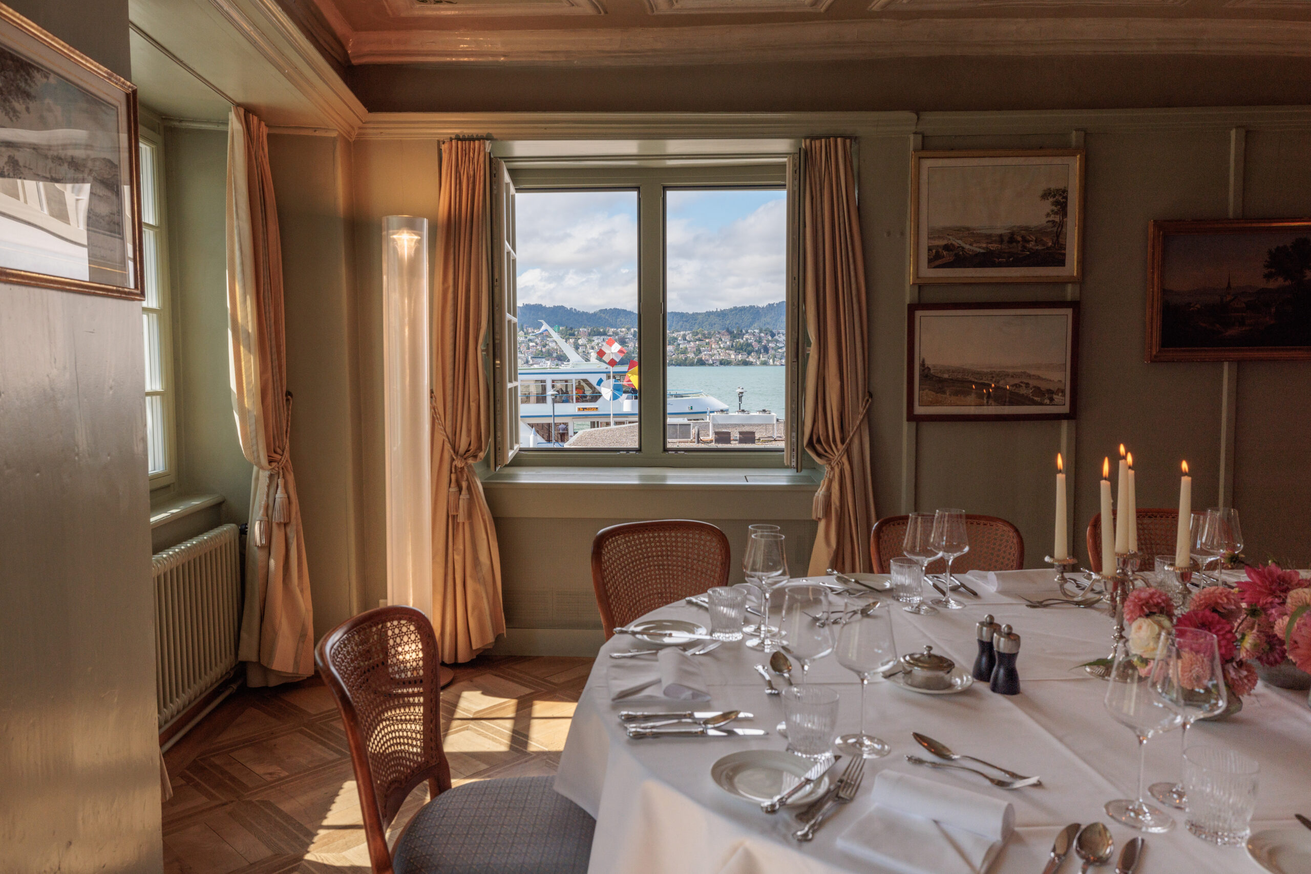 Ein runder, mit elegantem Geschirr und Kerzen gedeckter Esstisch befindet sich in einem sonnendurchfluteten Raum im Hotel Restaurant Sonne, mit Gemälden an den Wänden und einem Fenster, das den Blick auf den See mit Booten und fernen Hügeln freigibt.