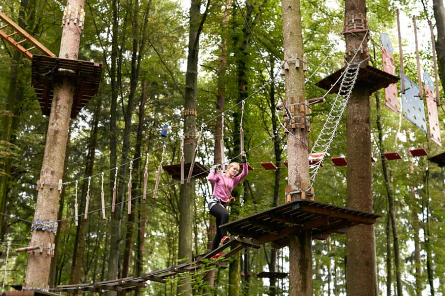 Eine Person, die eine rosa Jacke und einen Sicherheitsgurt trägt, bewegt sich auf einer Seilbrücke hoch zwischen den Bäumen in einem Seilpark, umgeben von Plattformen, Netzen und Kletterausrüstung in einem aufregenden Waldabenteuerpark.