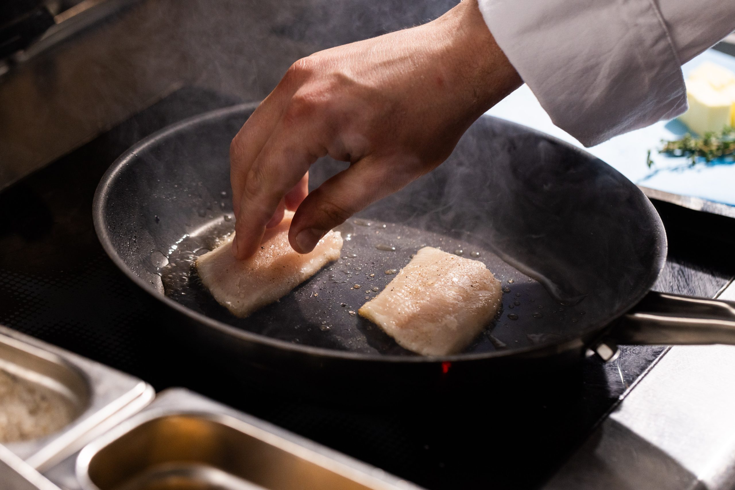 Die Hand eines Kochs legt ein Fischfilet in eine heiße, schwarze Pfanne auf dem Herd in der Küche Hotel Sonne, wo bereits ein weiteres Filet brät und Dampf aufsteigt.