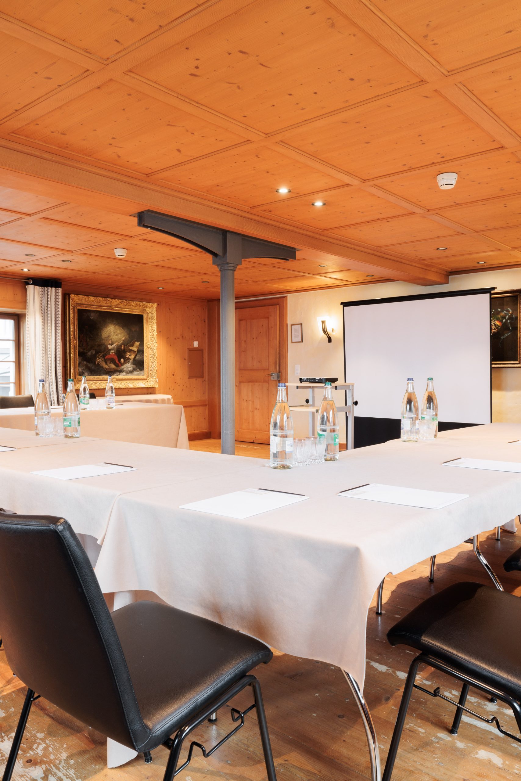 Ein Konferenzraum im Hotel Sonne mit hölzernen Wänden und Decken, eingerichtet für ein Seminar in U-Form mit schwarzen Stühlen, Wasserflaschen, Gläsern, Notizblöcken, einem Flipchart, einer Projektionsfläche und Gemälden an den Wänden.