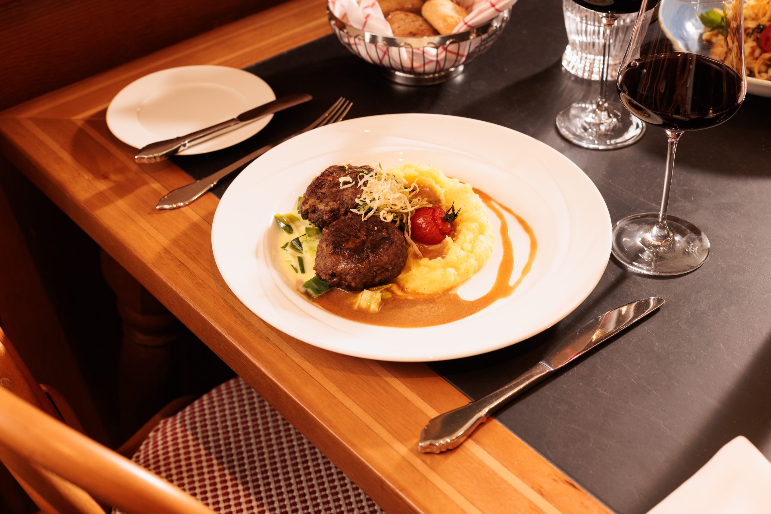 In der gemütlichen Gaststube im Hotel Sonne steht ein Teller mit Fleischklößchen mit Bratensoße, Kartoffelpüree, Gemüse und einer gebratenen Tomate, daneben ein Glas Rotwein, ein Brotkorb, Besteck und eine Serviette auf einem Holzstuhl.