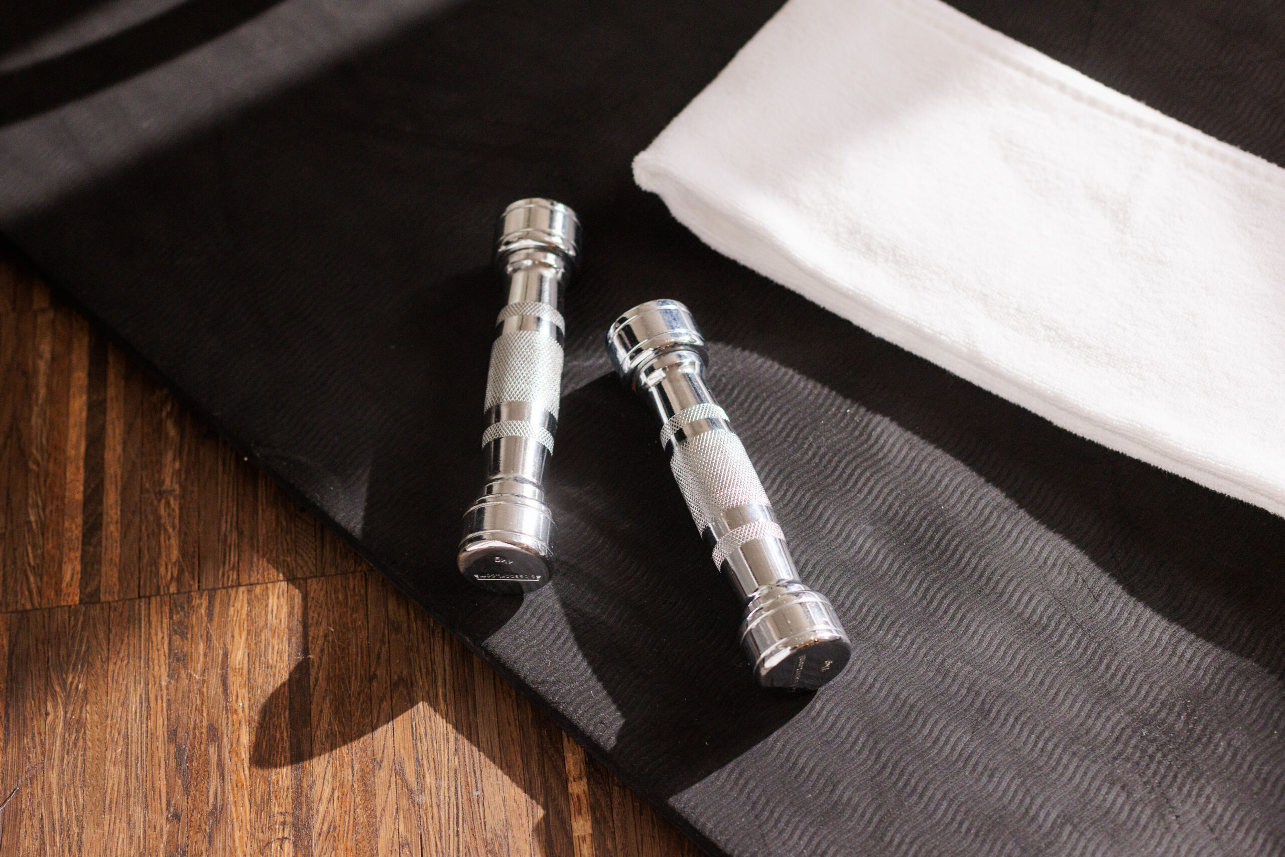 Zwei silberne Hanteln liegen auf einer schwarzen Trainingsmatte im Fitnessbereich, daneben liegt ein ordentlich gefaltetes weißes Handtuch. Das Sonnenlicht wirft Schatten auf den Holzboden und schafft eine ruhige Trainingsatmosphäre.