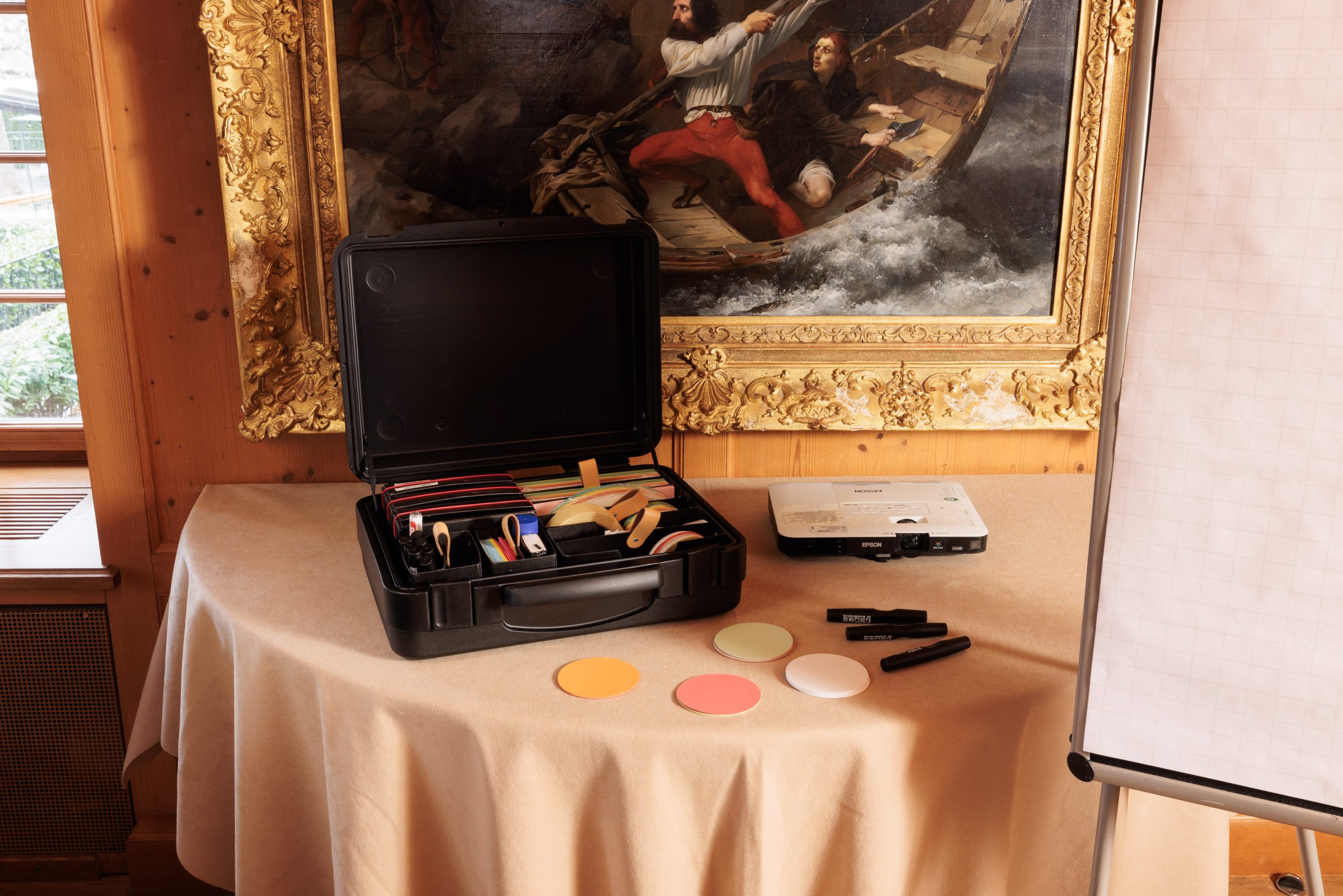 Ein runder Tisch im Hotel Sonne, aufgebaut für ein Seminar, mit einem schwarzen Koffer voller Büromaterial, einem Projektor, farbigen Papierkreisen und Markern. Im Hintergrund sind ein weißes Flipchart und ein großes gerahmtes Gemälde zu sehen.