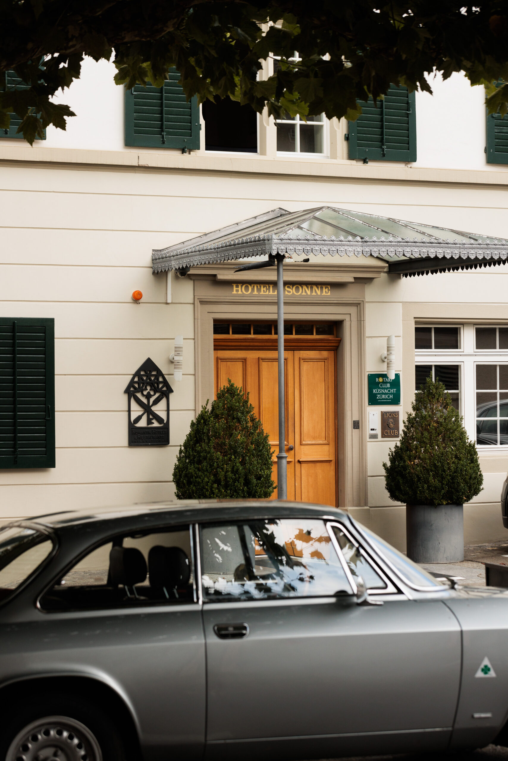 Ein silberner Oldtimer parkt vor einem cremefarbenen Boutiquehotel mit grünen Fensterläden, großen Topfpflanzen und einer Holztür unter einem Glasdach mit der Aufschrift HOTEL SONNE.