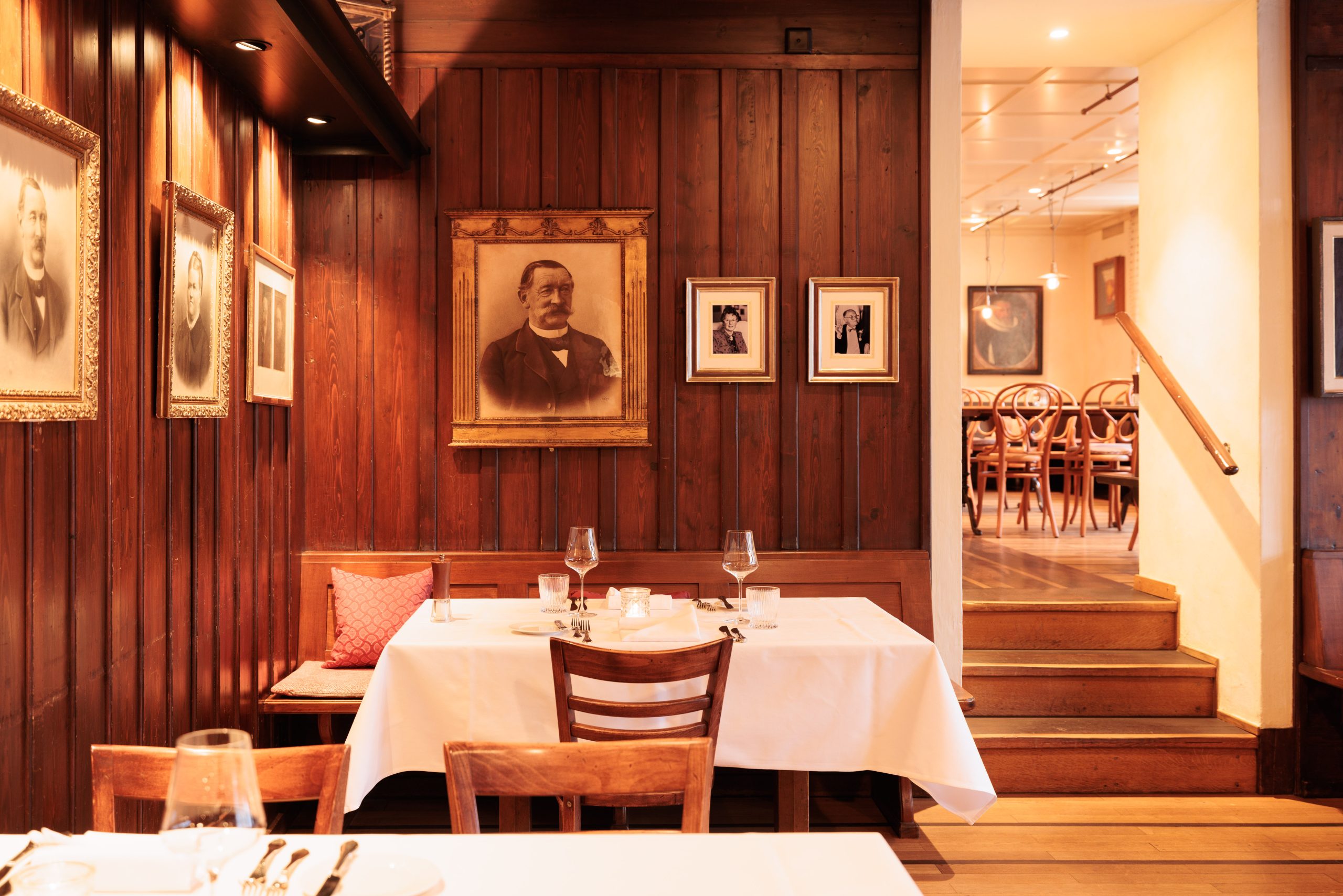 Ein gemütliches Interieur im Restaurant Holz mit Holzwänden, gerahmten Vintage-Porträts, einem gedeckten Tisch mit weißem Tuch, zwei Stühlen und einem Glas Wein; sanfte Beleuchtung spiegelt die traditionelle Kultur in einer warmen, einladenden Atmosphäre wider.