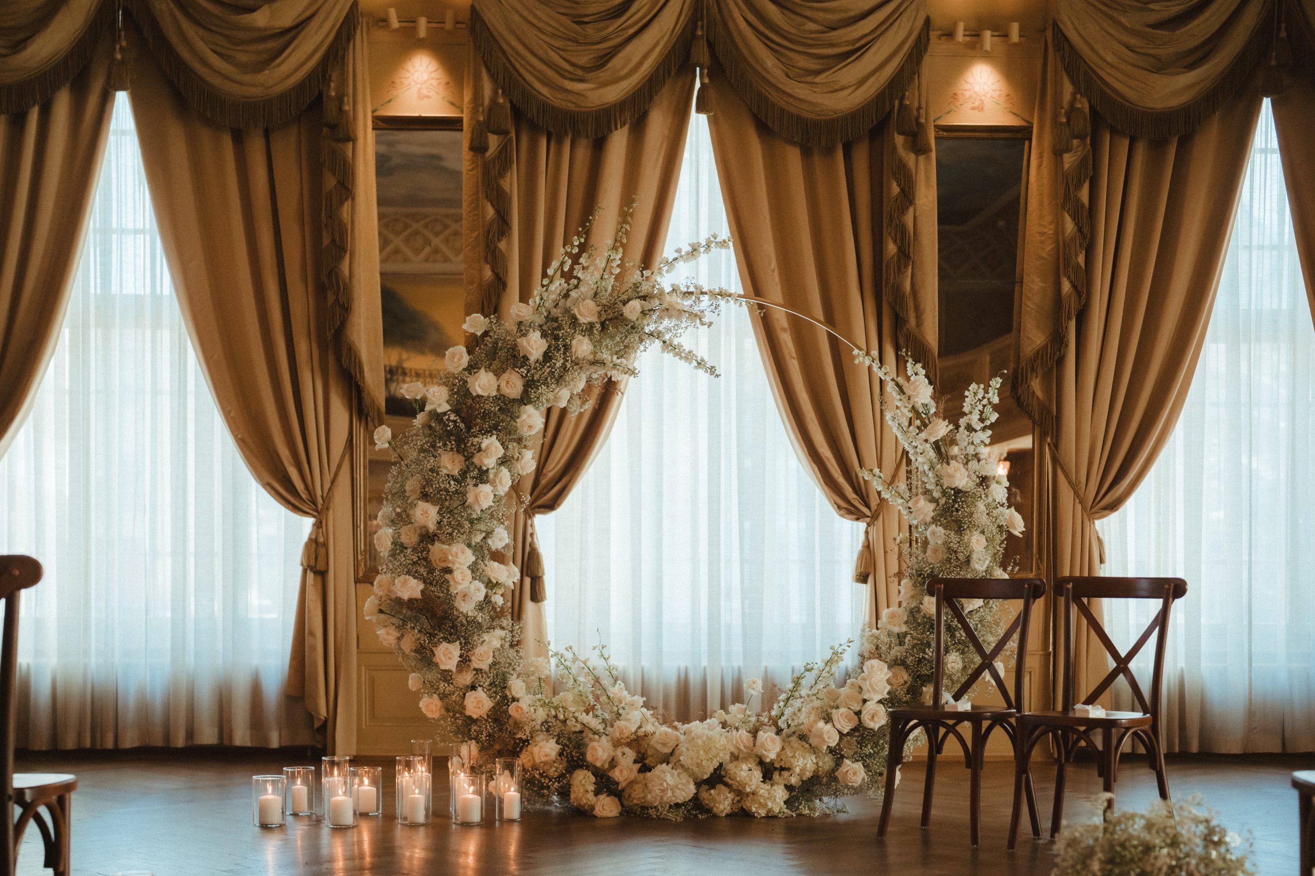 Elegantes Zimmer im Hotel Sonne mit hohen, goldenen Vorhängen und einem runden Blumenbogen aus weißen Blumen und Grünzeug. Zwei Holzstühle und Hochzeitsdeko-Kerzen sind vor dem Bogen angeordnet, während das Licht durch die Fenster hereinströmt.