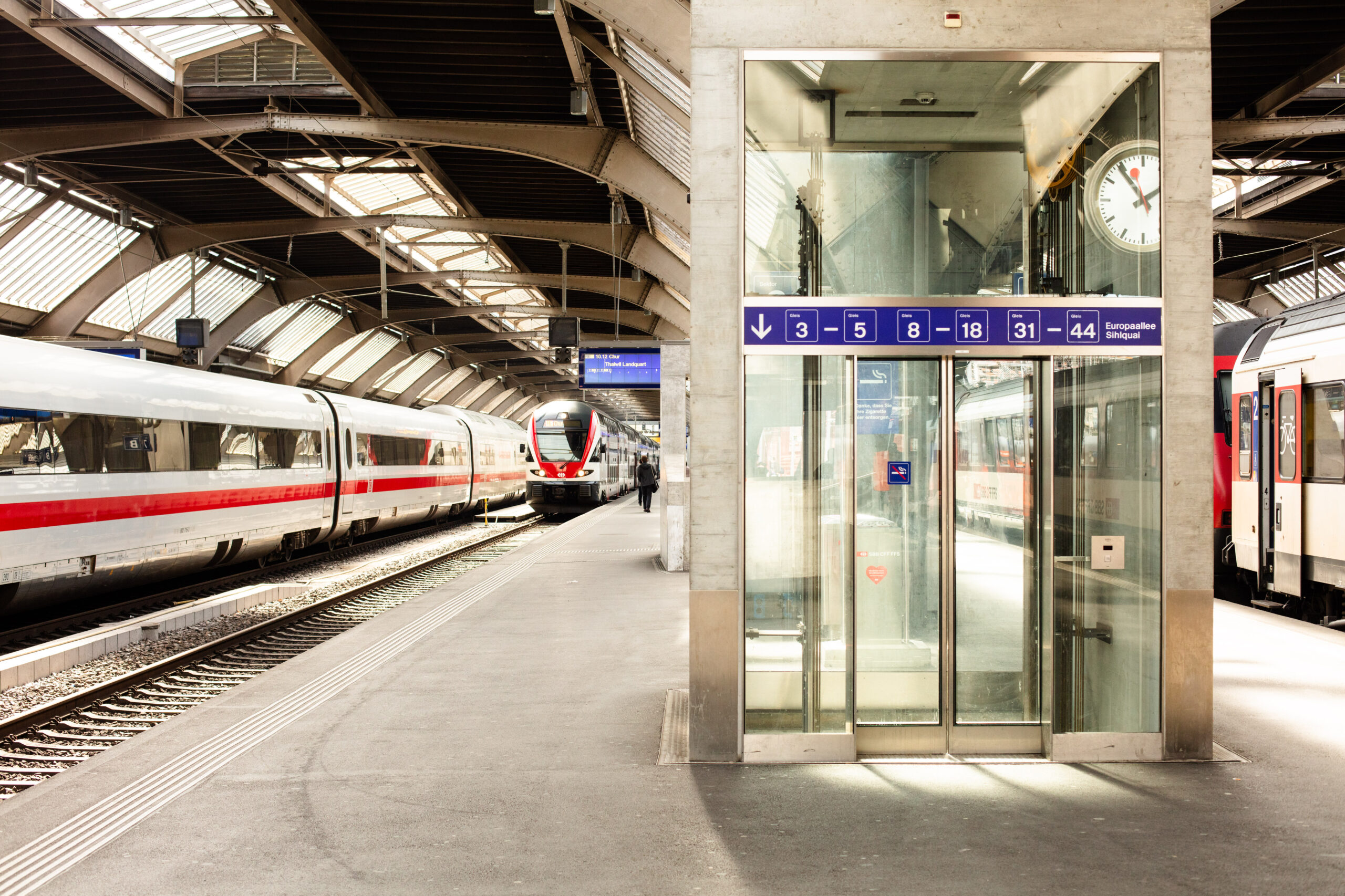 Ein moderner Bahnsteig in Zürich mit einem gläsernen Aufzug in der Mitte, digitalen Anzeigen über dem Kopf, einer großen Uhr und zwei weißen Zügen mit roten Streifen neben den Gleisen unter einem gewölbten Dach.