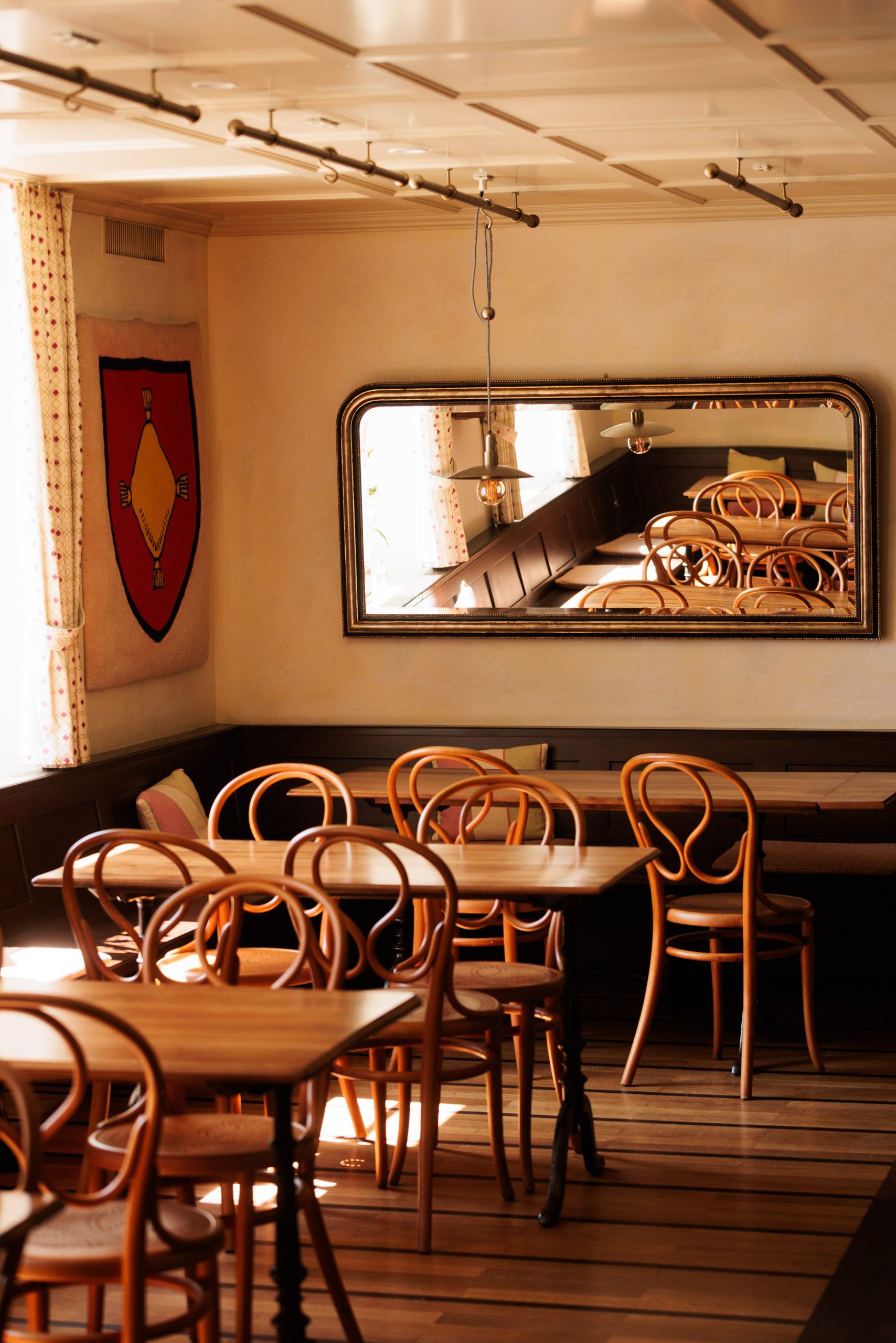 Die sonnenbeschienene, leere Gaststube im Hotel Sonne ist mit Holztischen und geschwungenen Stühlen ausgestattet; ein großer Wandspiegel reflektiert die Möbel und Fenster mit gemusterten Vorhängen und schafft eine warme und einladende Atmosphäre.