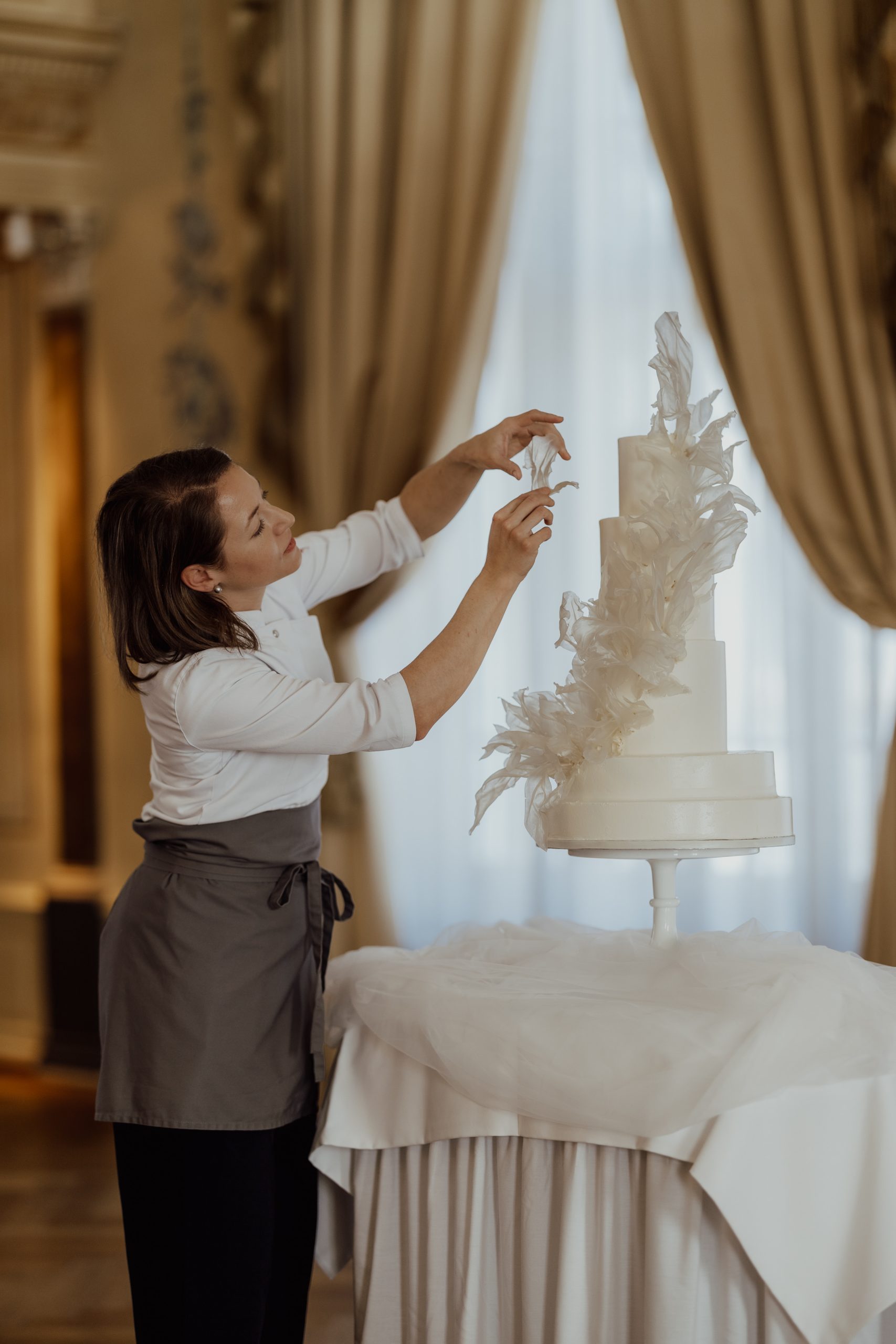 Eine Frau dekoriert eine hohe weiße Hochzeitstorte mit eleganten federartigen Details in einem verzierten Raum mit drapierten Fenstern, perfekt für eine stilvolle Hochzeit am See oder als Inspiration für Ihre Hochzeitslocation Zürichsee.