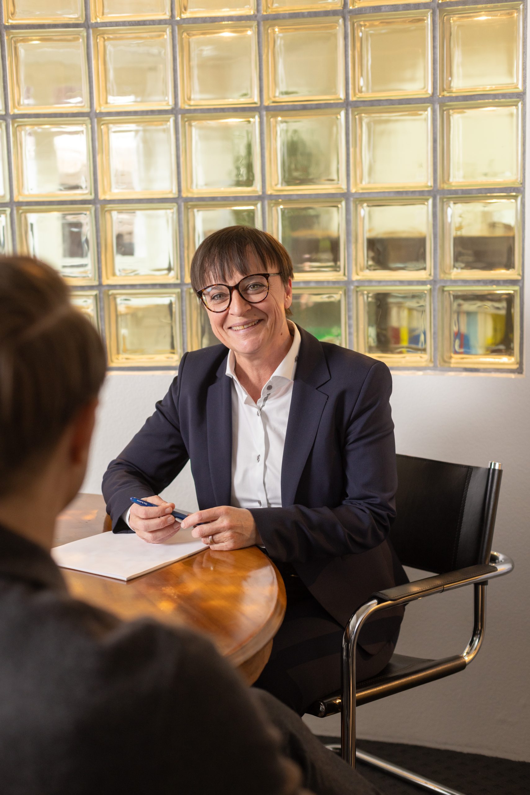 Eine Person im Anzug und mit Brille sitzt an einem Tisch, lächelt und hält einen Stift mit einem Notizblock vor sich, während sie einer anderen Person gegenübersitzt, die mit dem Rücken zur Kamera steht. Im Hintergrund ist eine Wand aus Glasbausteinen zu sehen.