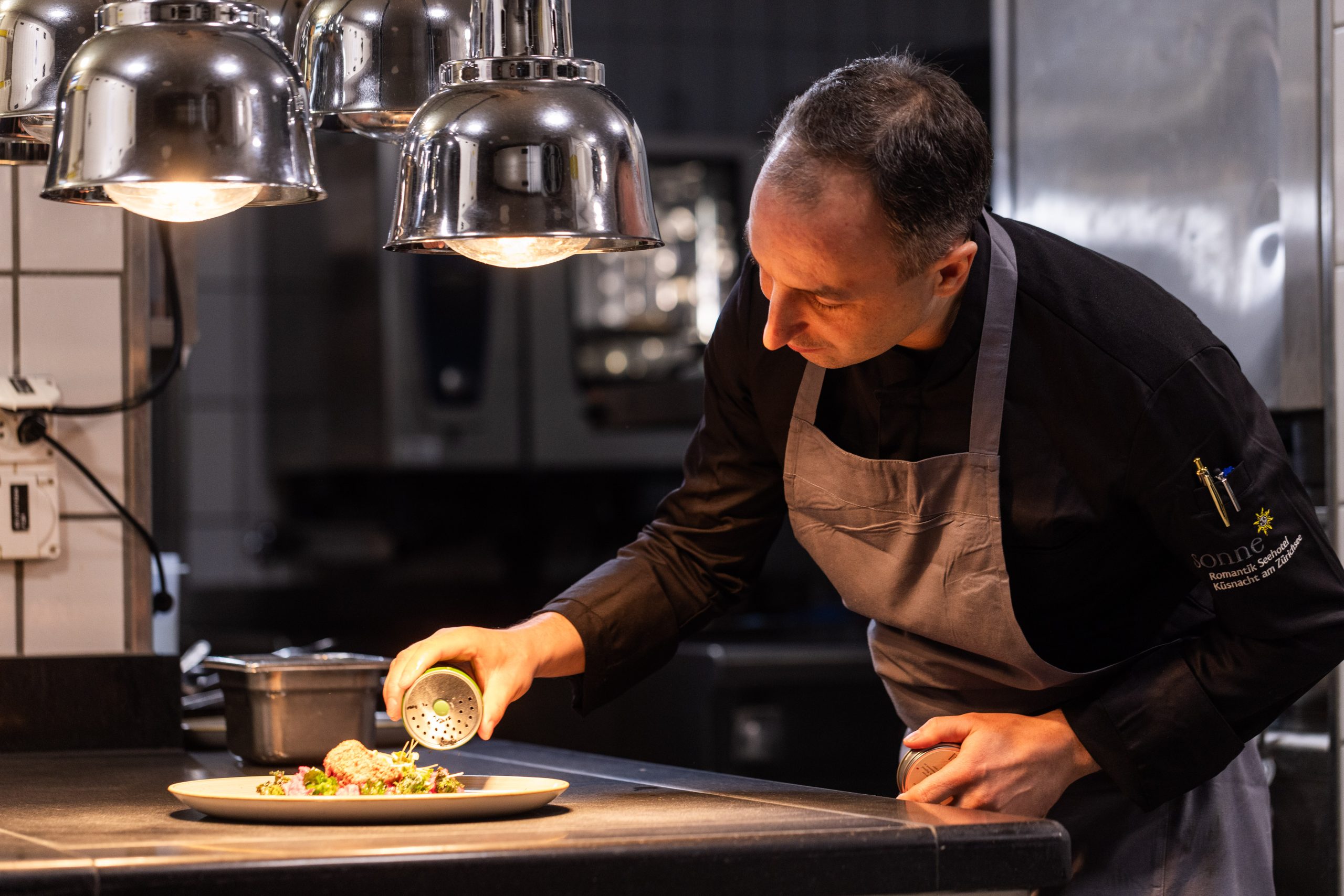 Ein Koch in schwarzer Uniform und grauer Schürze streut unter hellem Küchenlicht in der Profiküche des Küche Hotel Sonne sorgfältig die Gewürze auf ein Tellergericht.