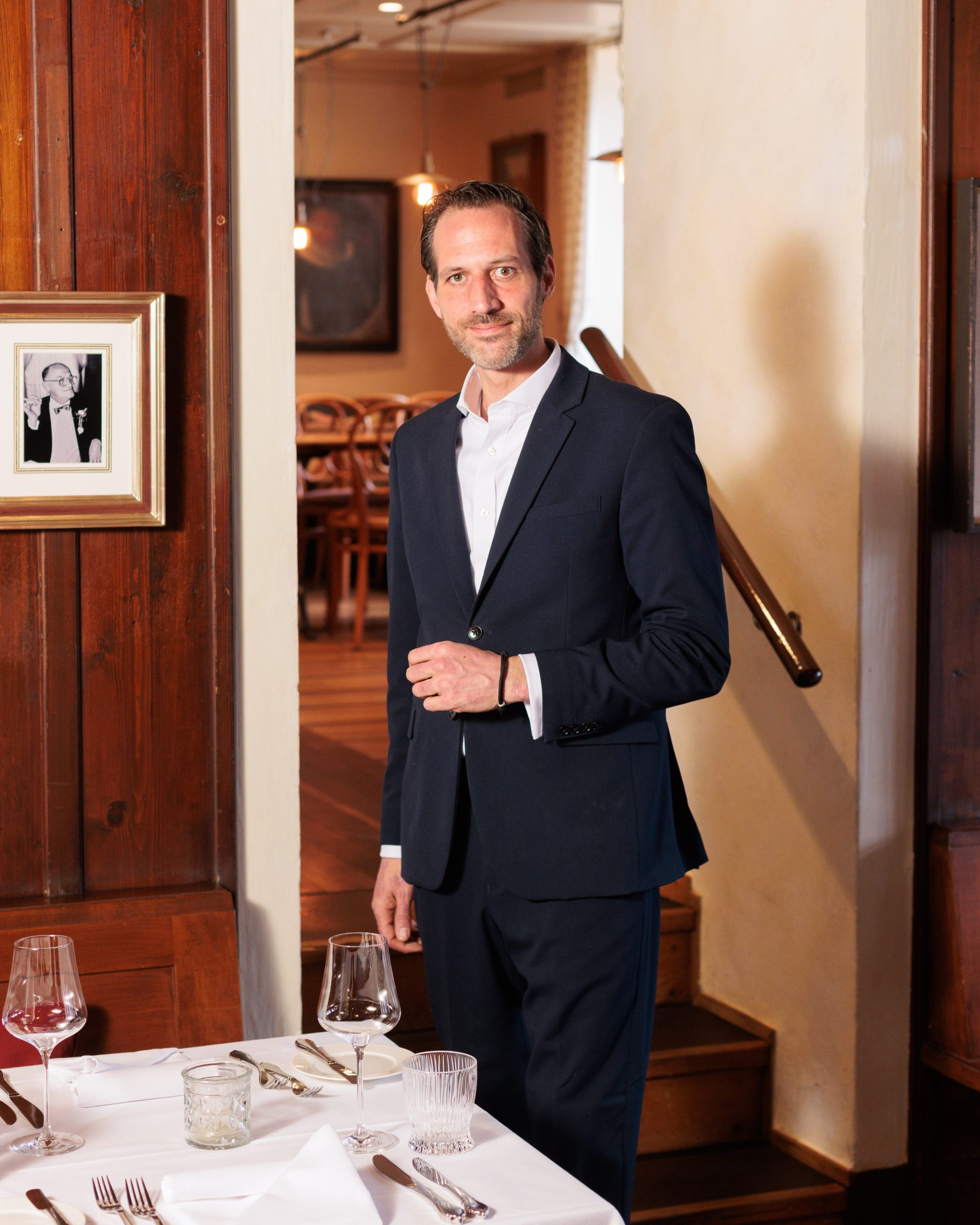 Ein Mann in einem dunklen Anzug steht in einem warm beleuchteten Restaurant neben einem ordentlich gedeckten Tisch und lächelt leicht. Weingläser und Geschirr sind auf dem weißen Tischtuch angeordnet. An der Holzwand hinter ihm hängt ein gerahmtes Schwarz-Weiß-Foto.
