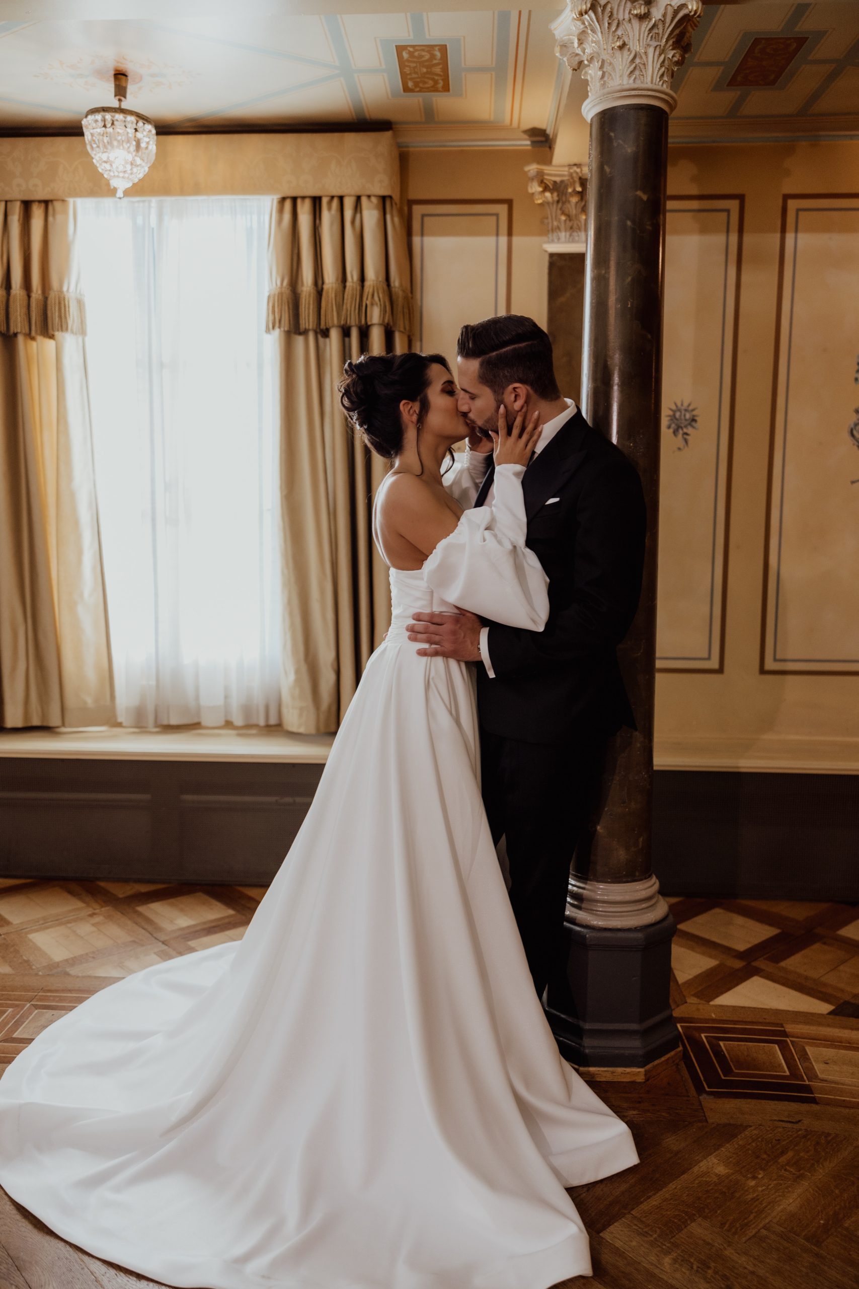 Eine Braut in einem klassischen Hochzeitskleid und ein Bräutigam in einem schwarzen Anzug umarmen sich und küssen sich in einem eleganten, sonnendurchfluteten Hotelzimmer mit goldenen Wänden, langen Vorhängen und einer Marmorsäule.