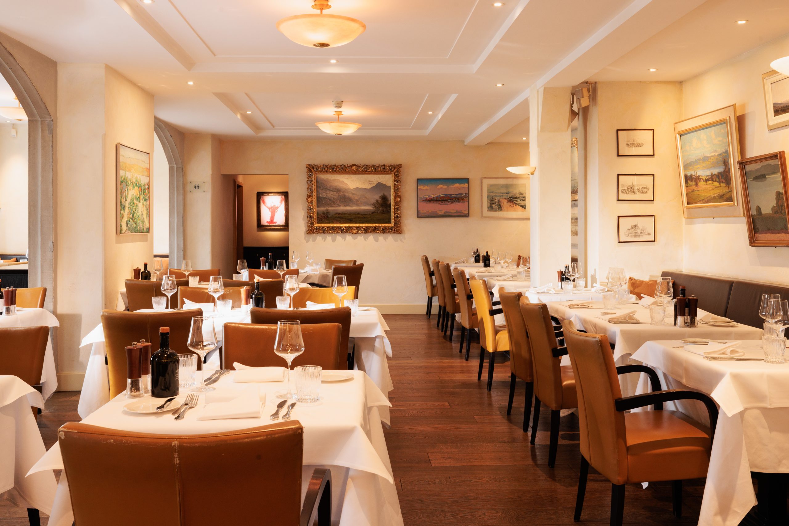 Elegantes Interieur im Restaurant Sonnengalerie mit ordentlich gedeckten Tischen mit weißen Tischtüchern, braunen Lederstühlen, Weingläsern und gerahmten Kunstwerken an cremefarbenen Wänden, die eine warme und einladende Atmosphäre schaffen.