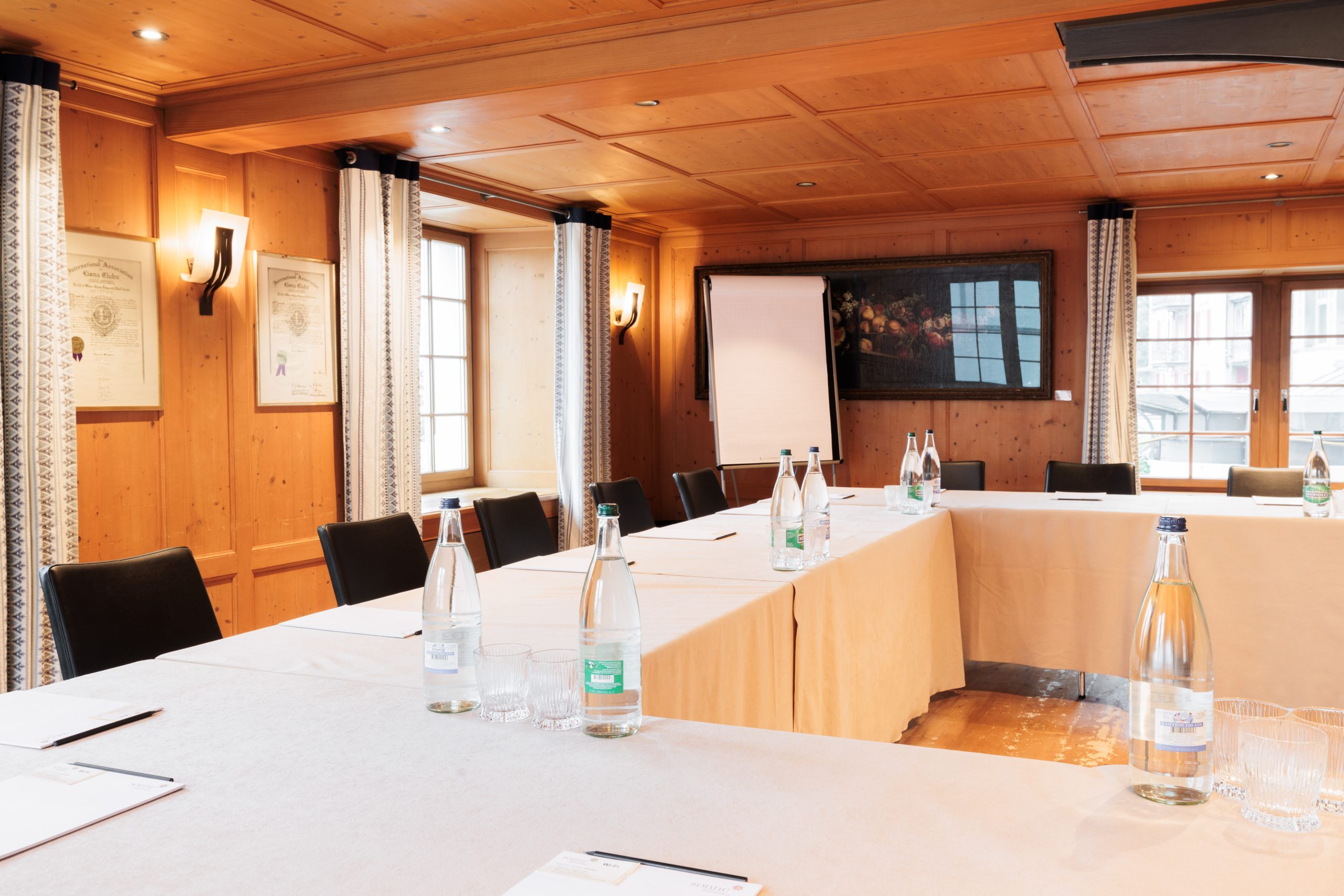 Ein heller Seminarraum im Hotel Sonne ist mit Holzwänden und -decken, U-förmigen Tischen, schwarzen Stühlen, Wasserflaschen, Gläsern, Notizblöcken, einem Flipchart, gerahmten Zertifikaten und großen Fenstern mit Vorhängen ausgestattet.