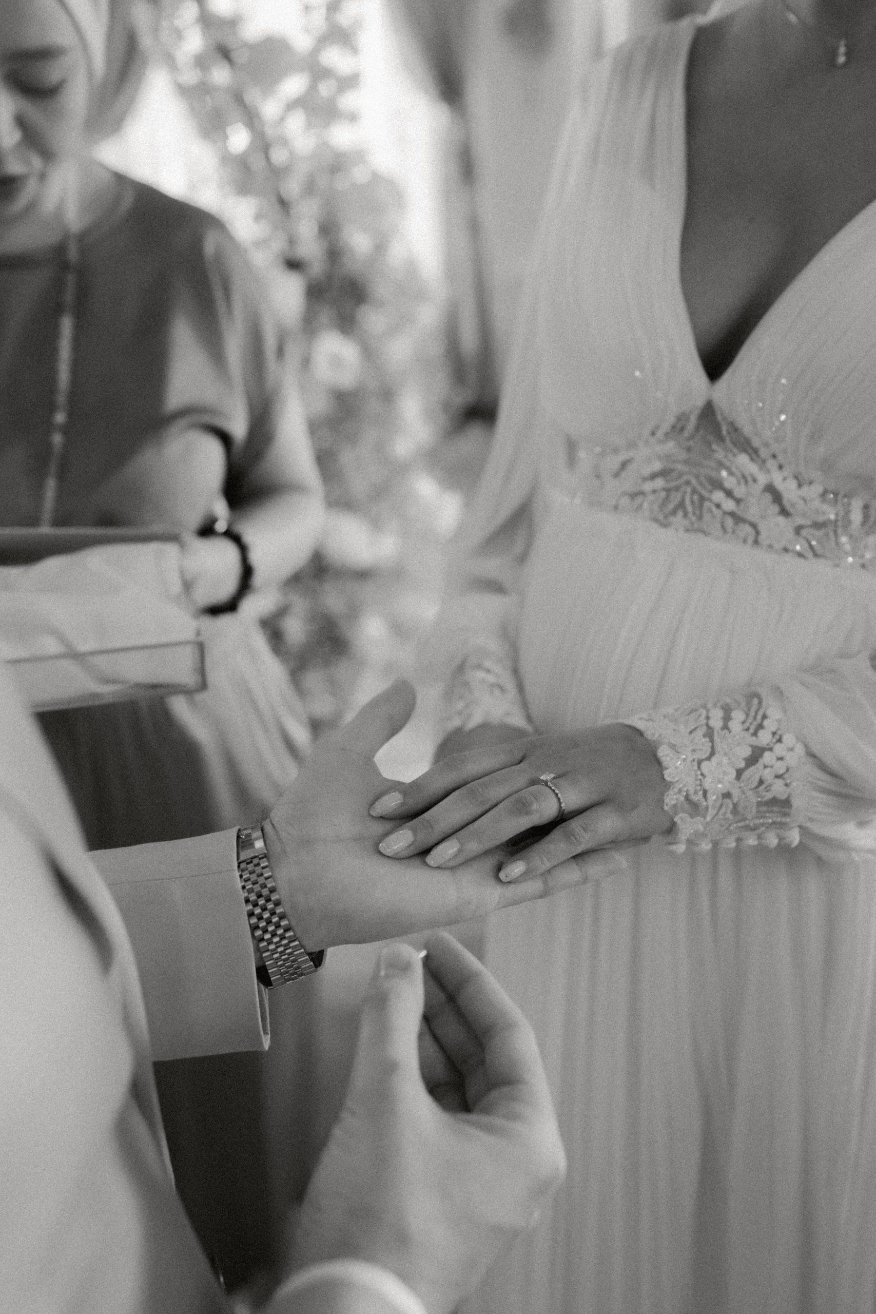 Eine Schwarz-Weiß-Nahaufnahme einer Traunung Hochzeit zeigt eine Person, die einer anderen einen Ring an den Finger steckt, die Braut in einem langärmeligen Kleid, während jemand in der Nähe eine Schachtel hält - vielleicht für Ringe oder sogar eine Rolex.