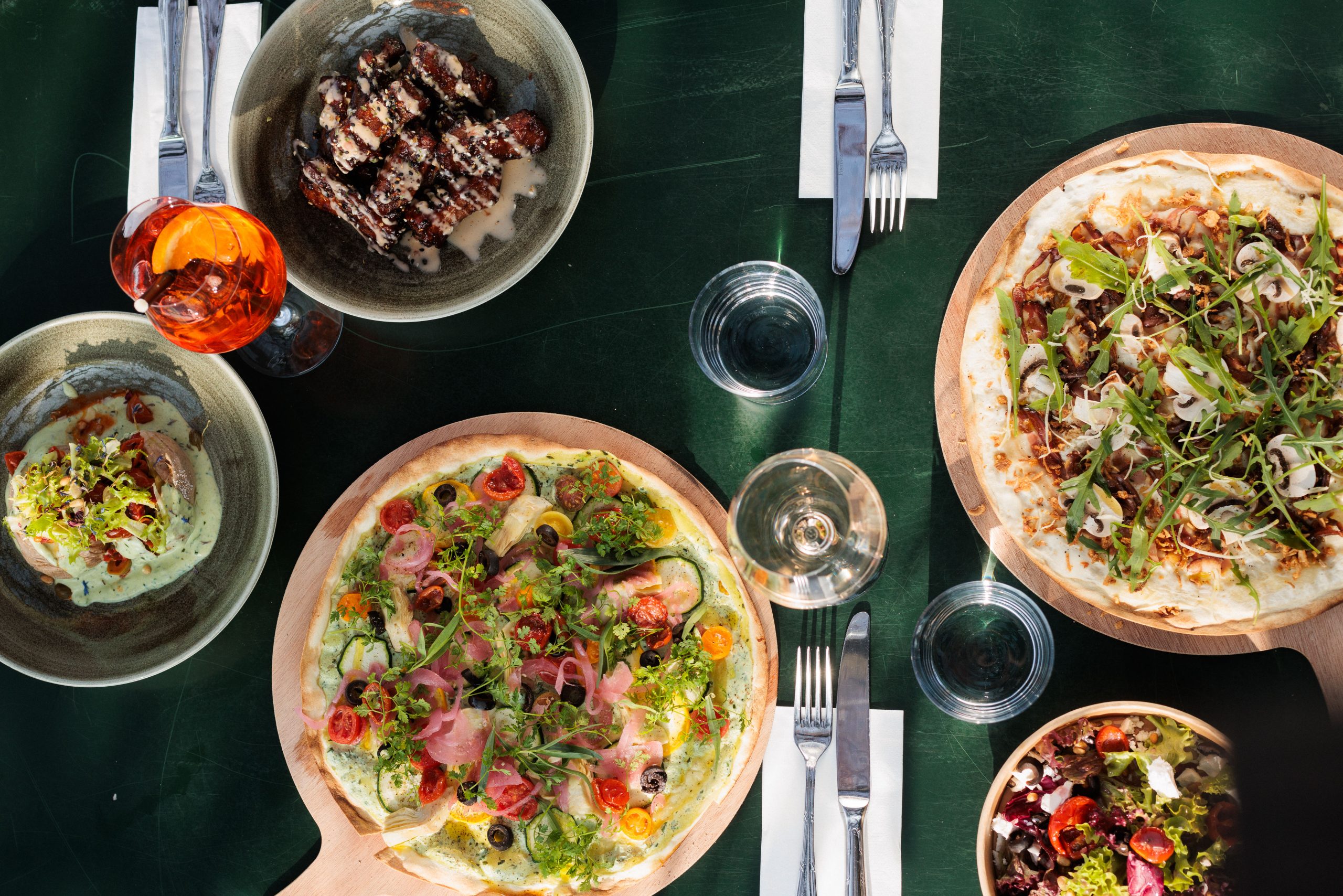 Draufsicht auf einen Tisch in einem Biergarten mit zwei Pizzen, die mit Grünzeug belegt sind, einem Salat, Hummus mit Beilage, einer Schüssel Fleisch mit Soße, Gläsern mit Wasser und einem Orangencocktail auf einer dunkelgrünen Fläche.