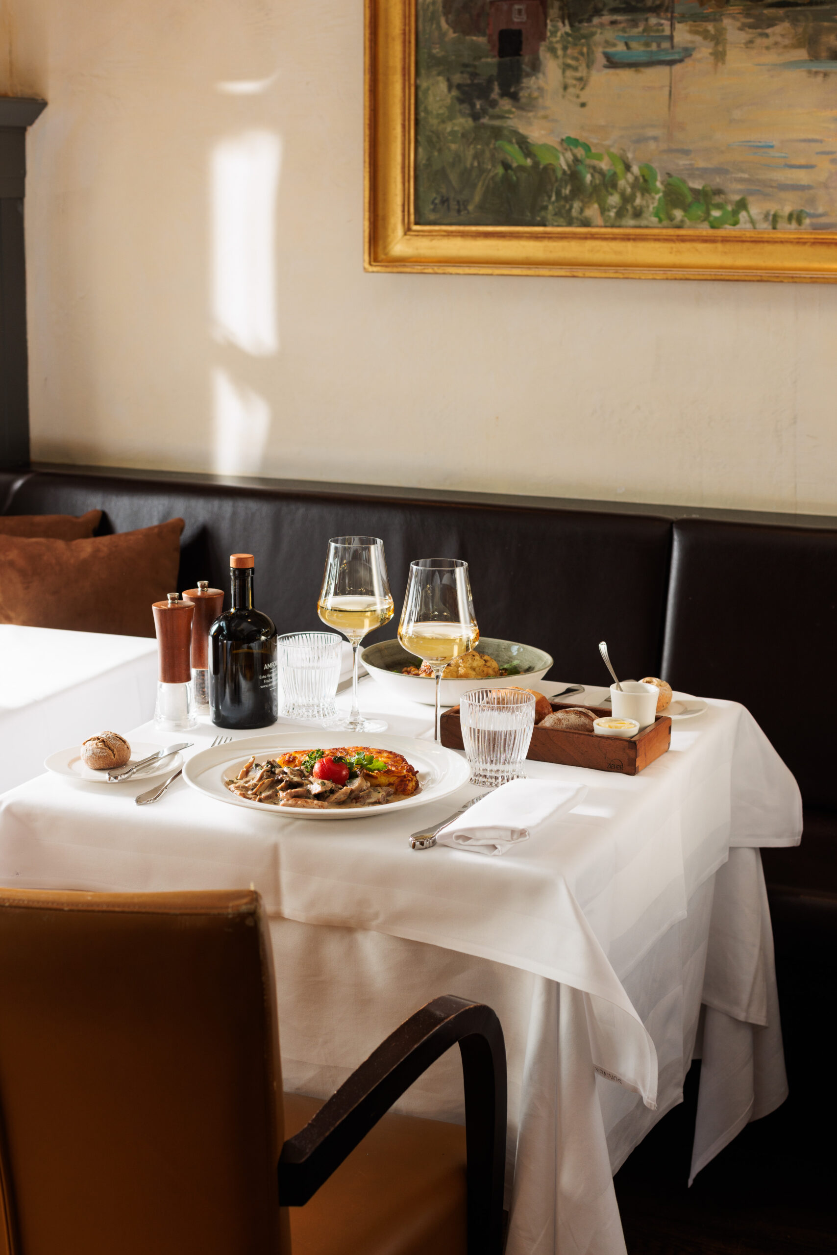 Ein sonnendurchfluteter Restauranttisch für zwei Personen in der Sonnengalerie, gedeckt mit weißem Tuch, Tellern mit Speisen, Weingläsern, Brot und Gewürzen. An der Wand über der dunklen Sitzbank hängt ein Gemälde.