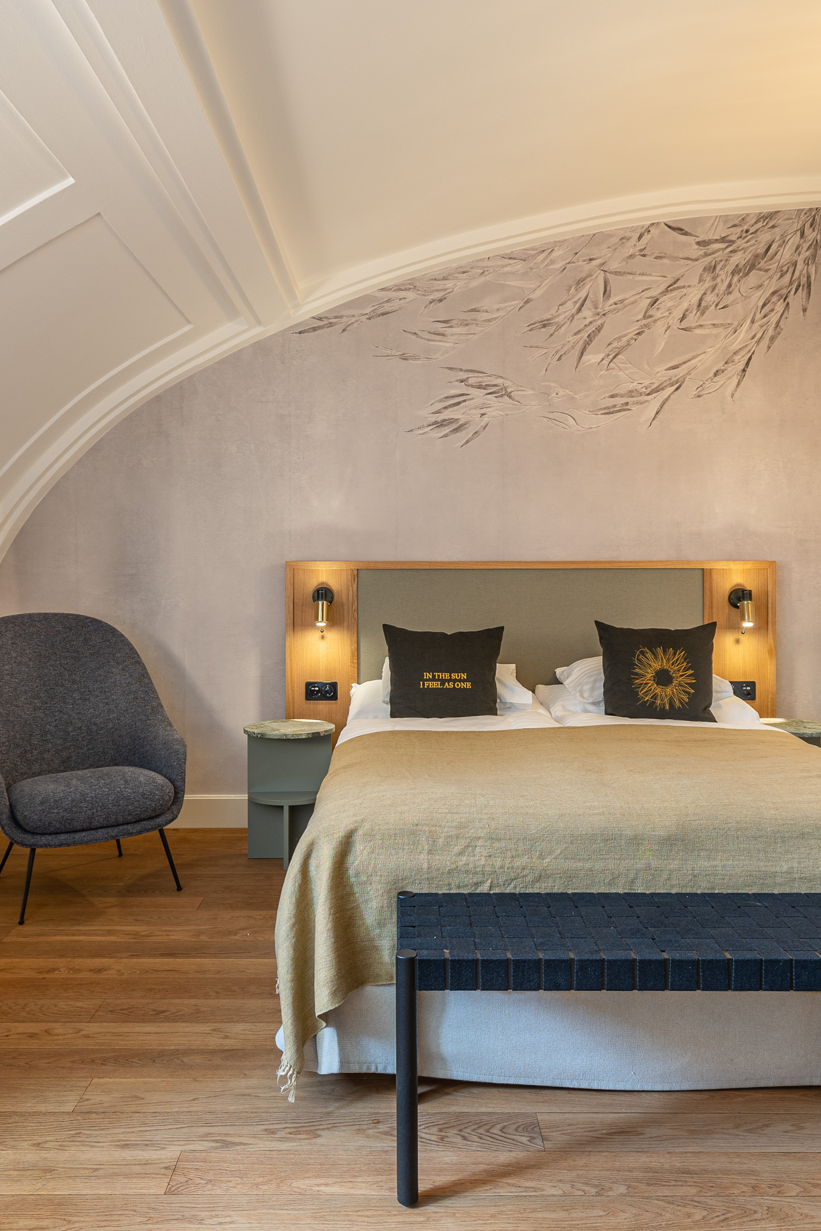 Es erwartet Sie ein gemütliches Hotelzimmer am Zürichsee mit einem Doppelbett, dekorativen Kissen, einer grünen Überwurfdecke, Wandleuchten, einem grauen Sessel, einer schwarzen Bank, hellem Holzboden und dezenter Kunst über dem Bett.