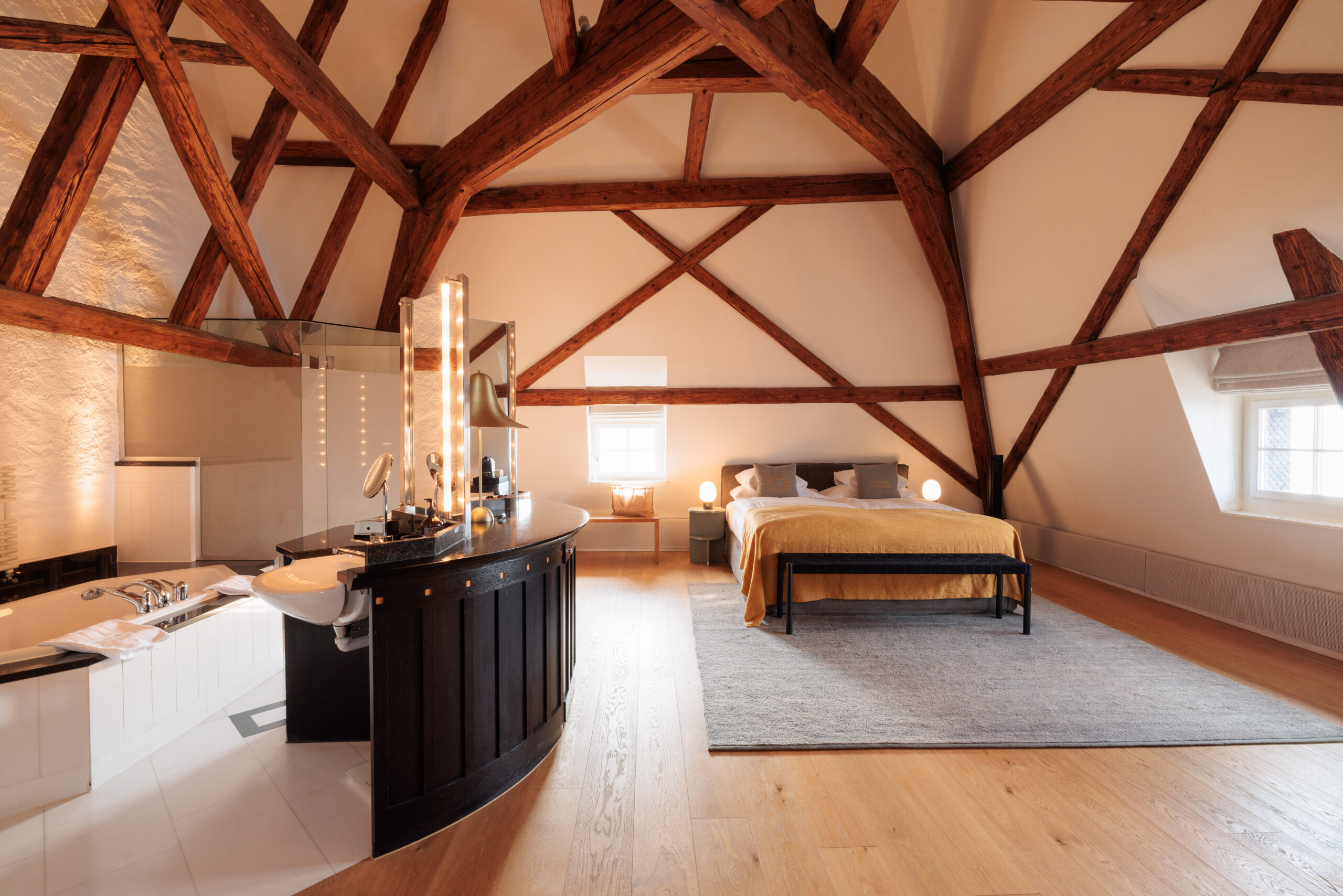 Das geräumige Loft-Zimmer im Hotel Sonne Küsnacht verfügt über freiliegende Holzbalken, ein grosses Bett mit gelber Bettwäsche, einen grauen Vorleger und ein offenes Badezimmer mit moderner Beleuchtung. Natürliches Licht erfüllt den Raum und sorgt für einen entspannten Aufenthalt.
