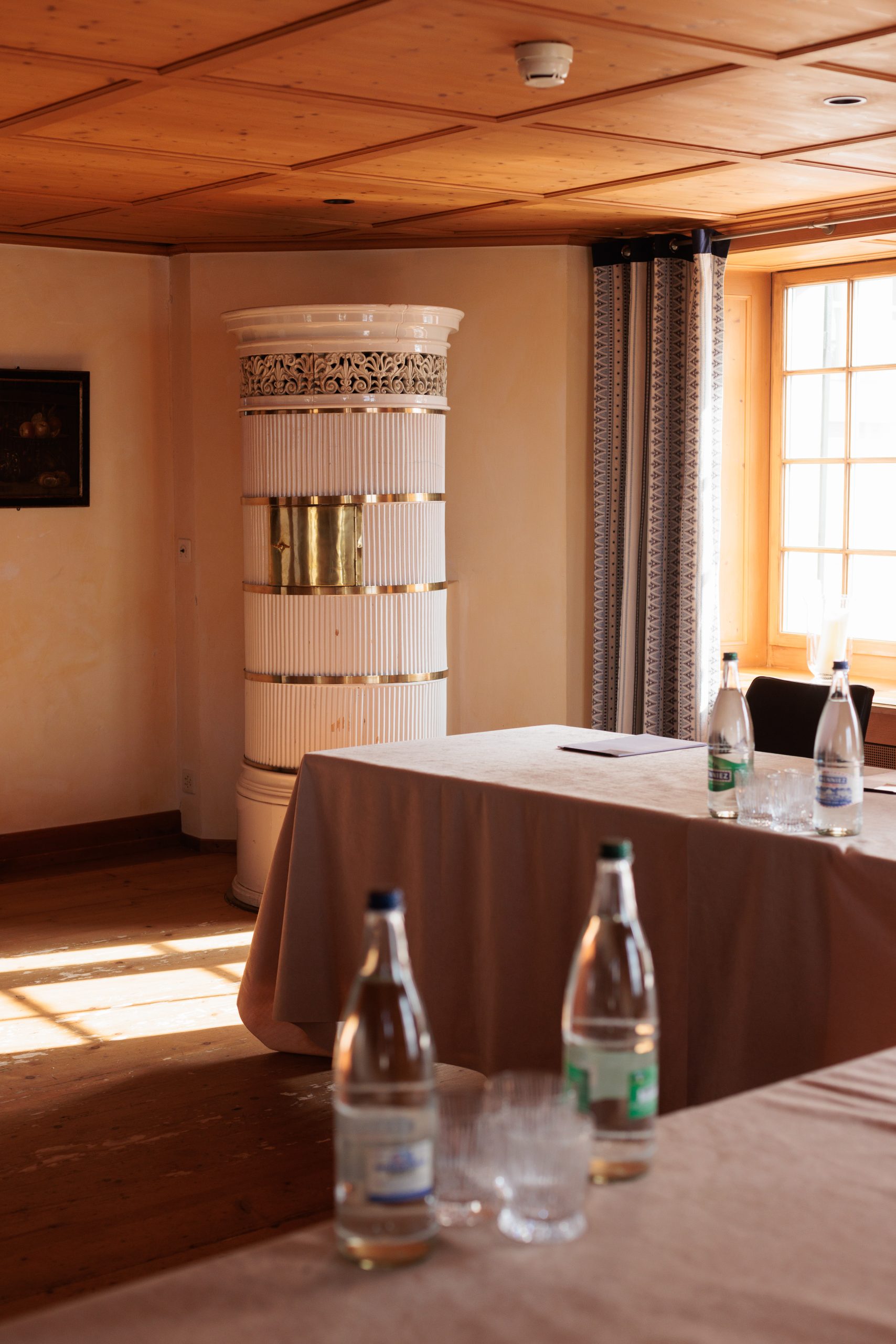 Ein sonnendurchfluteter Raum im Hotel Sonne mit Holzfußboden und -decke, mit Seminartischen, die mit beigem Stoff bezogen sind, Wasserflaschen, Gläsern, einem Fenster mit Vorhängen und einem weißen, dekorativen Kachelofen in der Ecke.