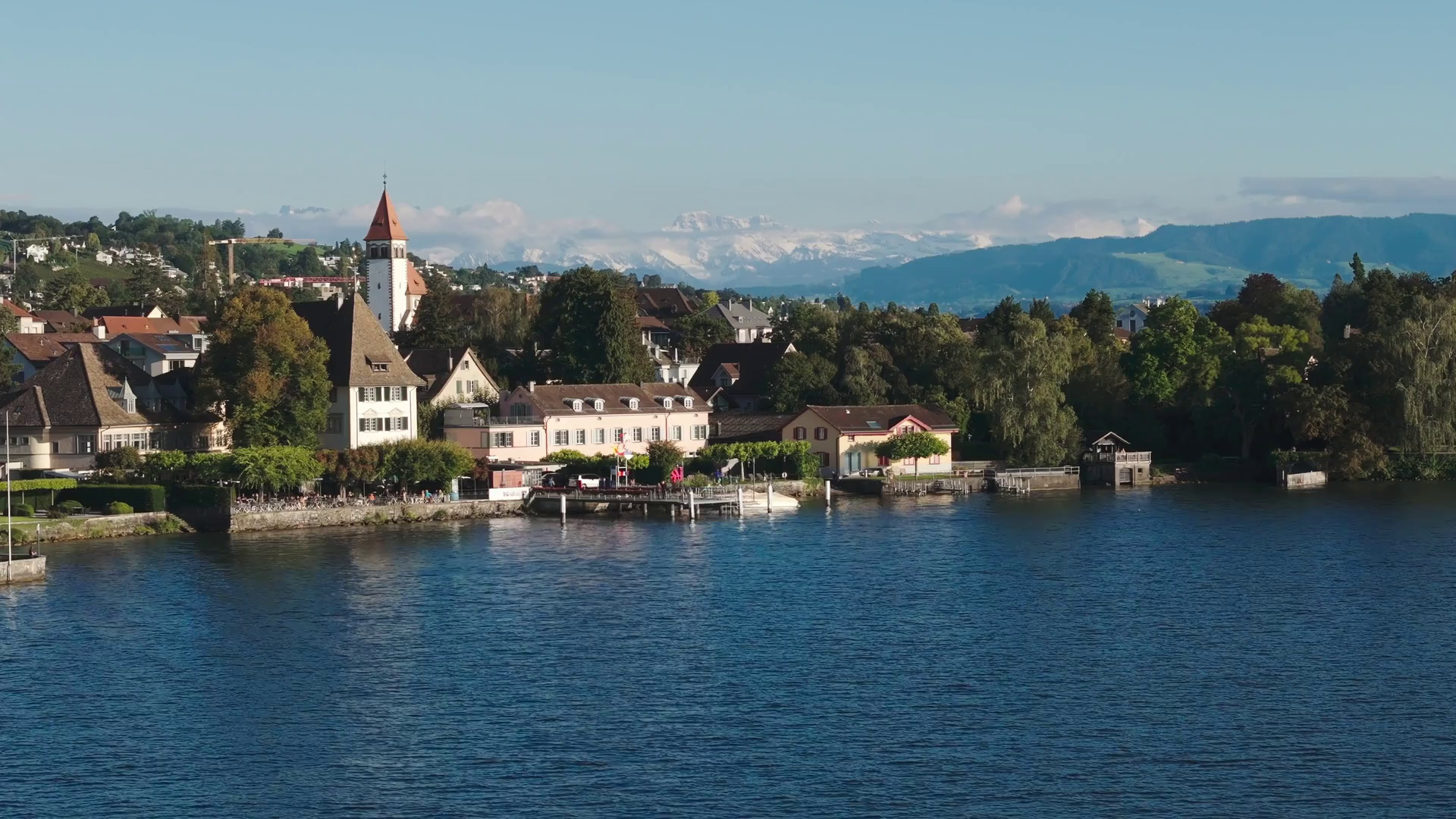 Ein friedliches Dorf am See mit traditionellen Gebäuden und dem Hotel Sonne in der Nähe eines Kirchturms, umgeben von Bäumen. Im Hintergrund sind die schneebedeckten Berge und der blaue Himmel über dem ruhigen Wasser zu sehen.