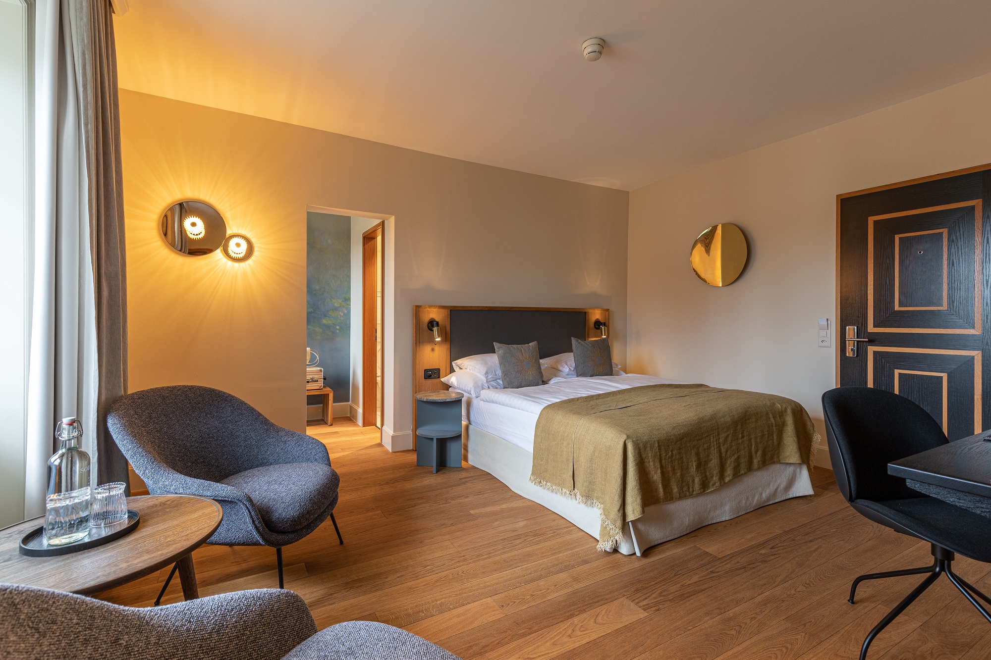 Ein modernes Boutiquehotelzimmer mit einem Doppelbett, brauner Decke, zwei grauen Stühlen und einem runden Tisch mit Wasserflaschen und Gläsern, Holzfußboden, stilvollen Wandleuchten und einer Tür, die in ein anderes Zimmer führt.