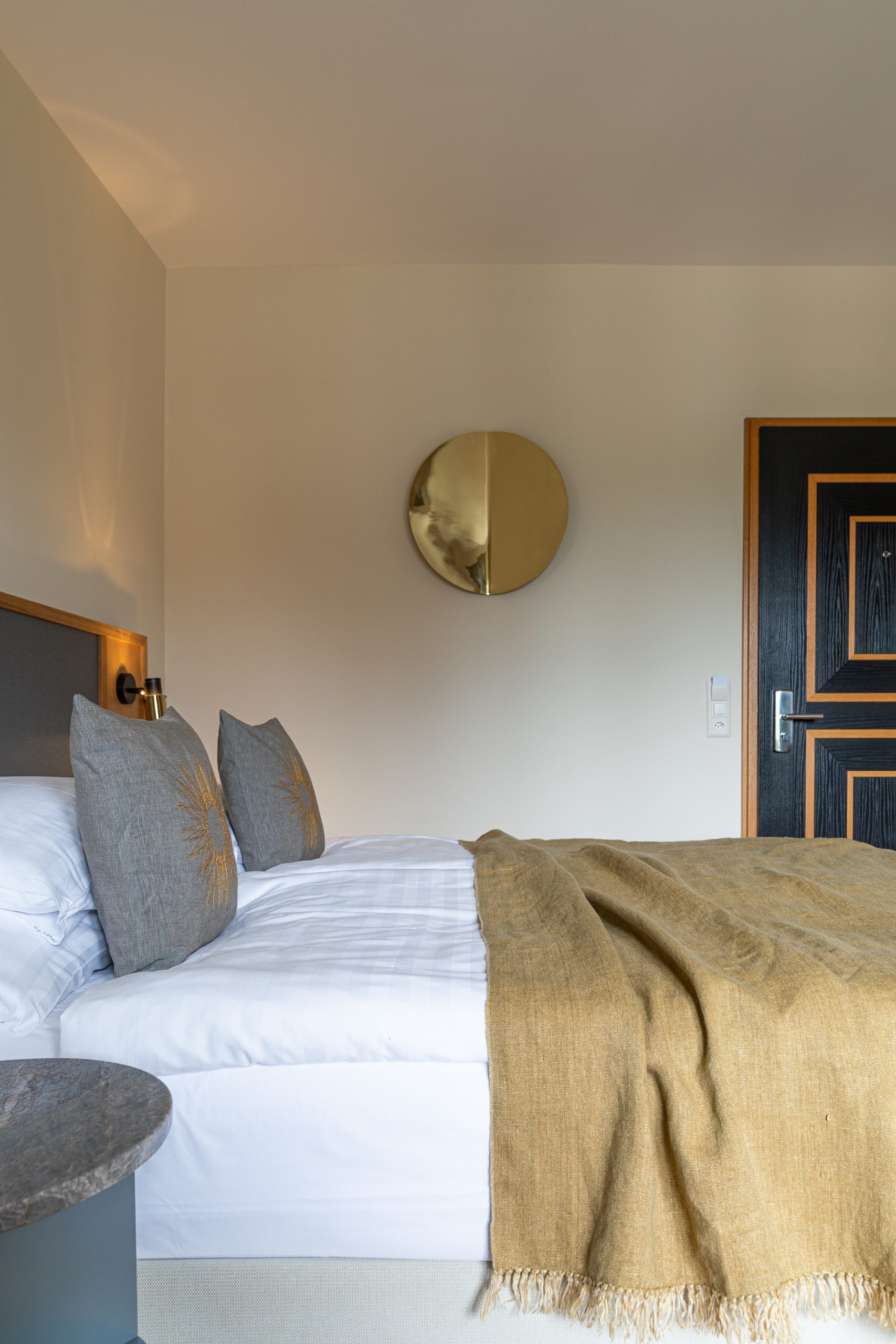 Ein ordentlich gemachtes Bett mit weißer Bettwäsche, zwei grauen Zierkissen und einer hellbraunen Decke steht neben einem runden Wandspiegel und einer dunklen Holztür in diesem modernen, minimalistischen Schlafzimmer im Stil eines Boutique-Hotels.