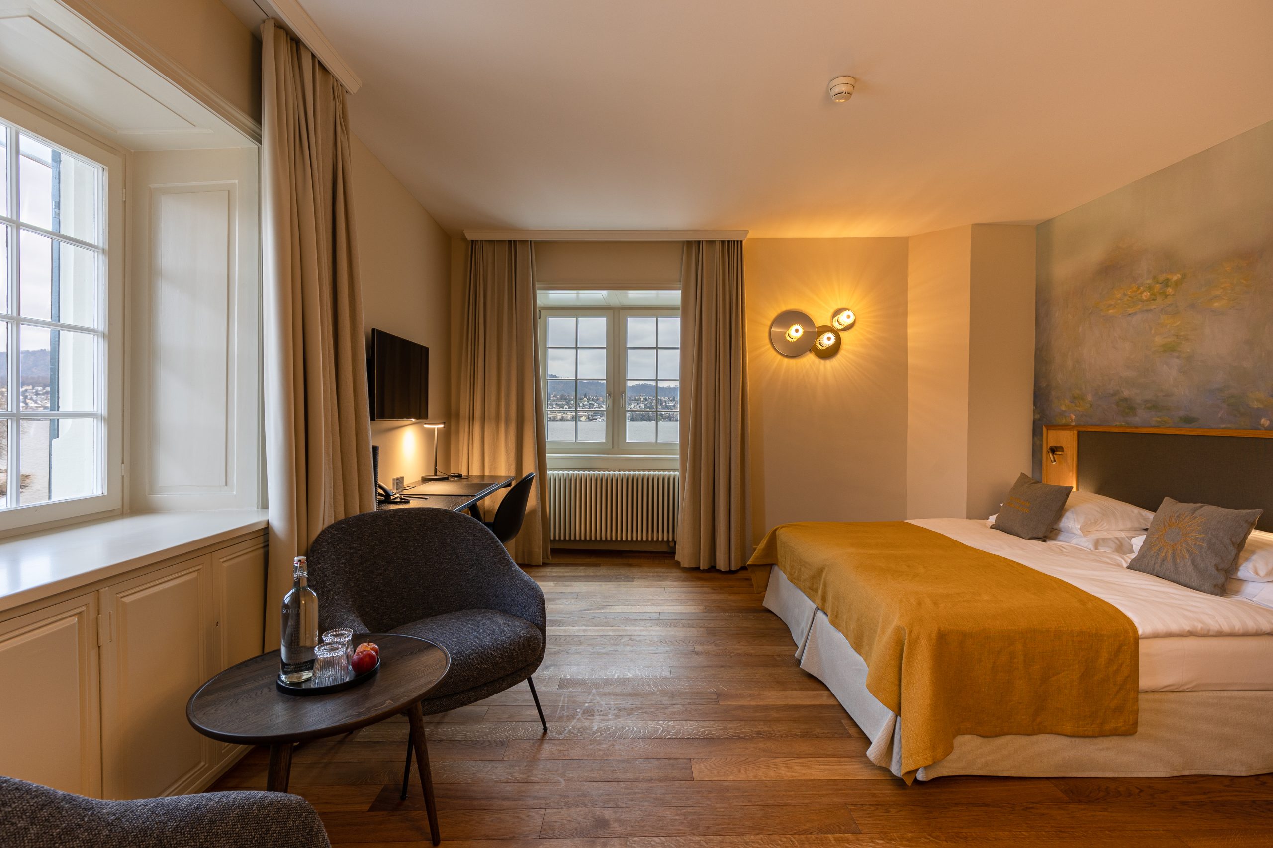 Ein gemütliches Boutique-Hotelzimmer mit einem Doppelbett mit gelber Bettwäsche, zwei grauen Stühlen um einen kleinen Tisch mit einer Flasche Wasser und einem Glas, einem Schreibtisch mit Computer, Holzboden und großen Fenstern, durch die Tageslicht fällt.