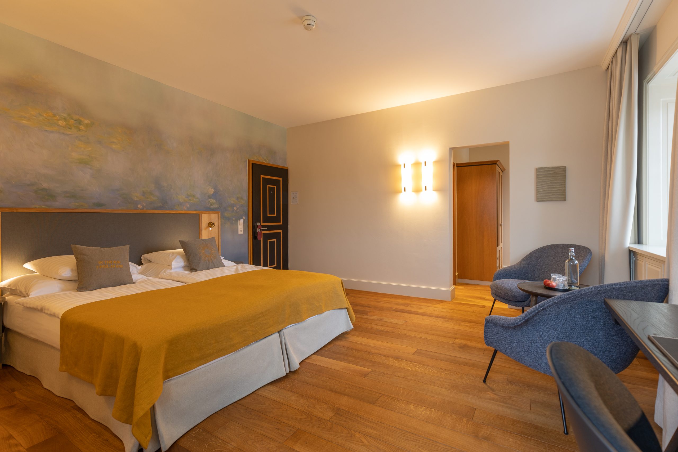 Ein gemütliches Boutique-Hotelzimmer mit einem Kingsize-Bett, das mit gelben Decken bedeckt ist, zwei blauen Stühlen, einem kleinen Tisch mit Wasser und Gläsern, Holzböden, einer Wandmalerei über dem Bett und warmer Beleuchtung.