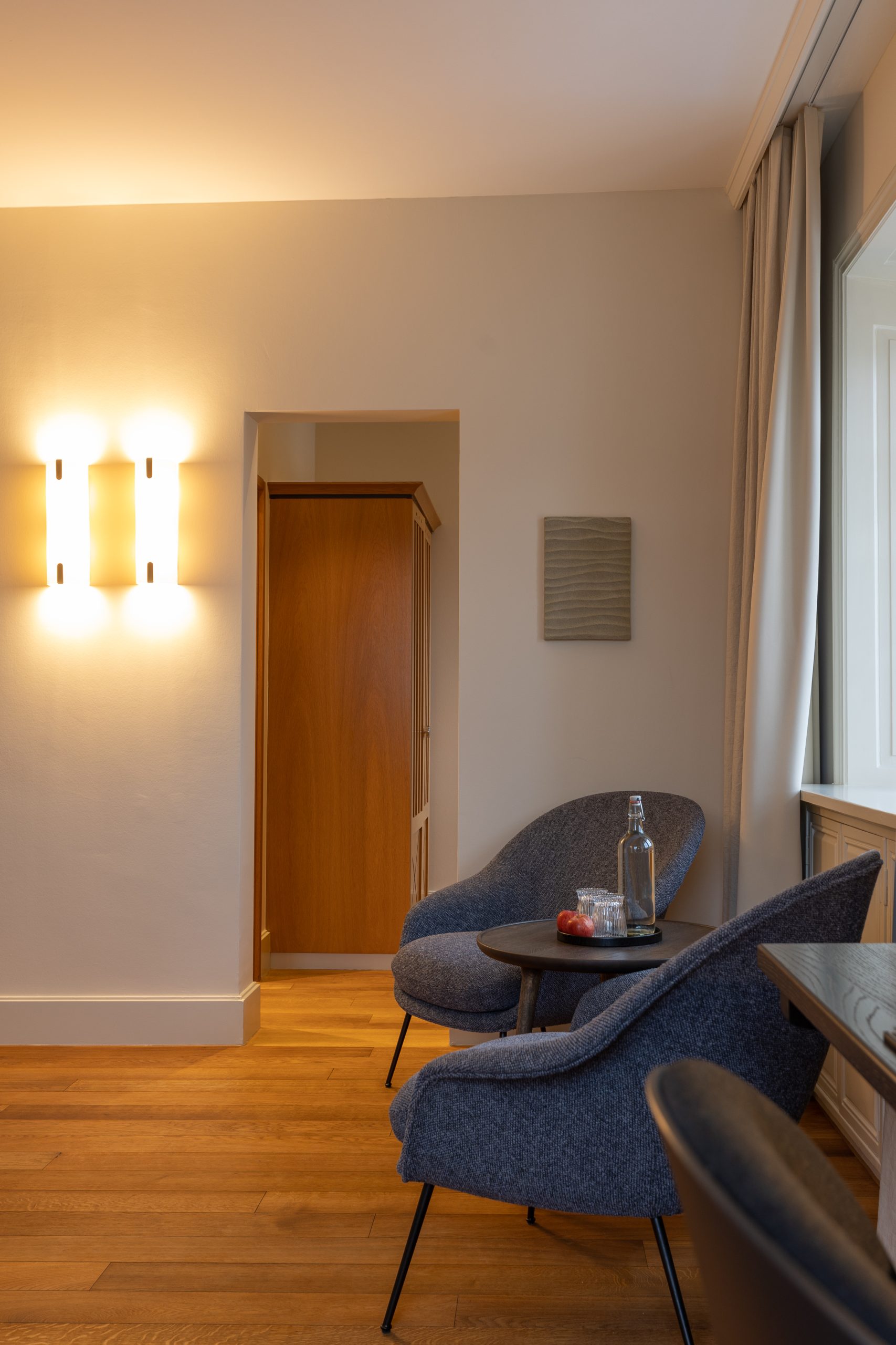 Eine gemütliche Boutiquehotel-Sitzecke mit zwei blau gepolsterten Stühlen und einem kleinen runden Tisch mit einer Glasflasche und einem Glas auf einem Holzboden in der Nähe eines Fensters und einer sanft beleuchteten Wand.