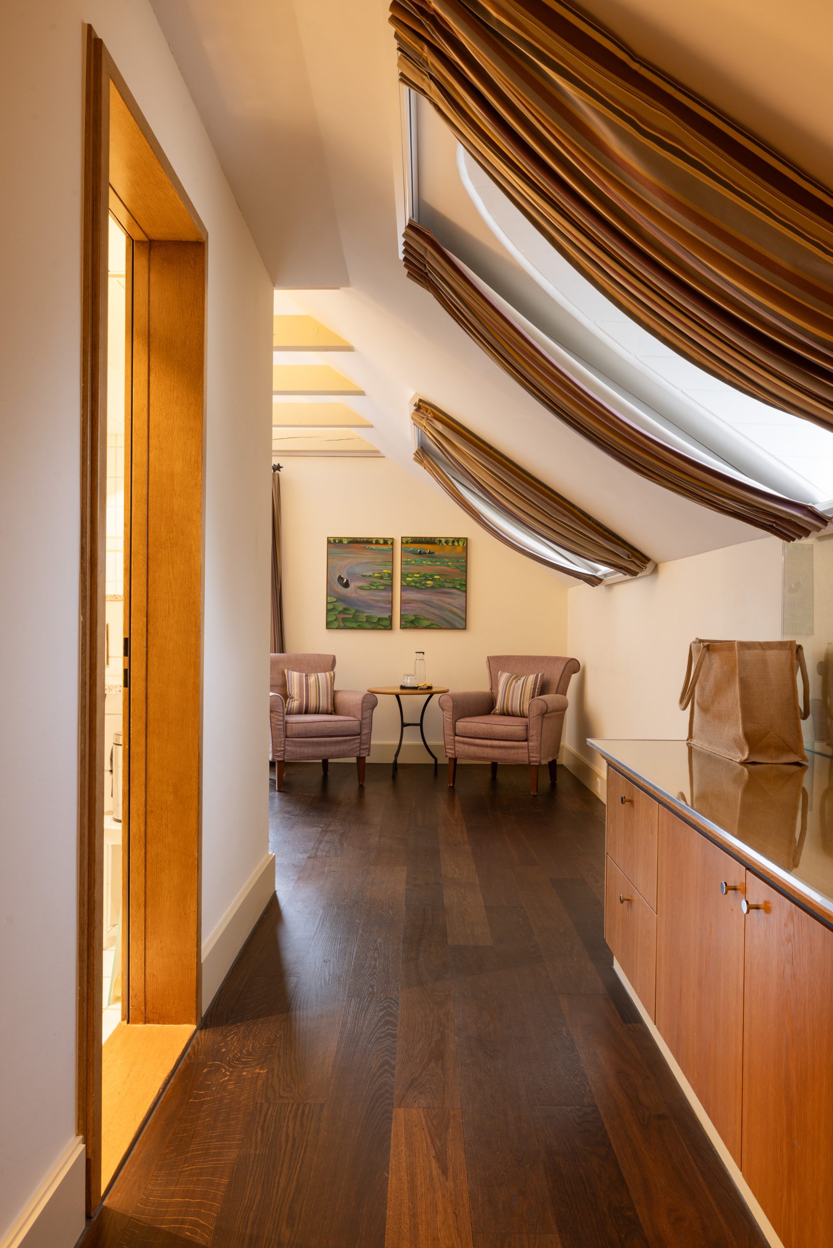 Die gemütliche Sitzecke in diesem Boutiquehotel ist mit zwei gestreiften Sesseln und einem kleinen Tisch ausgestattet, eingerahmt von schrägen Decken mit gestreiften Vorhängen, großen Dachfenstern, Holzböden und eleganten Schränken.