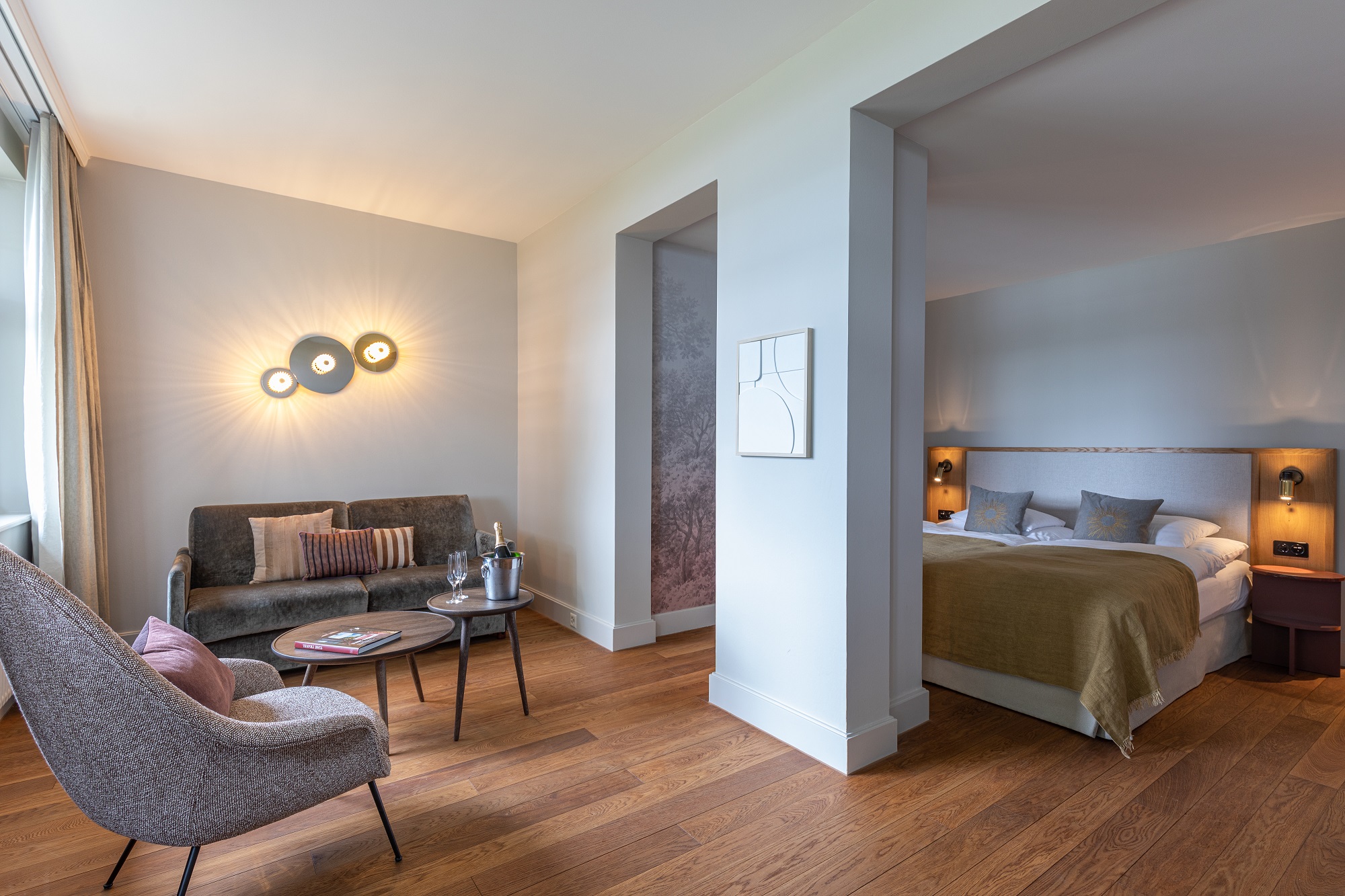 Ein modernes Boutiquehotelzimmer mit Holzboden, einem Bett mit grüner Bettwäsche, einem grauen Sofa, einem runden Couchtisch und einem grauen Sessel. Sanfte Beleuchtung und neutrale Wände schaffen eine gemütliche Atmosphäre.