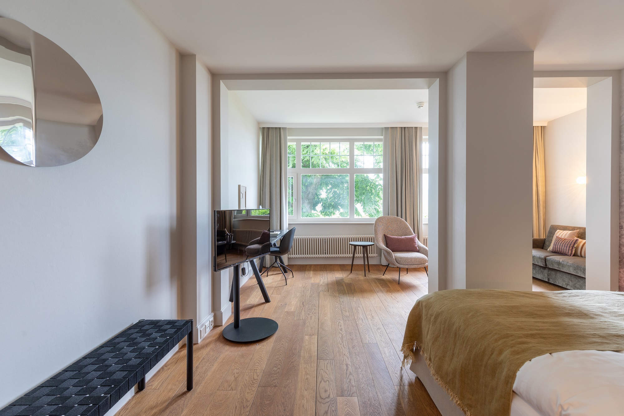 Helles, modernes Boutiquehotel-Zimmer mit Holzboden, einem Bett mit goldener Decke, Flachbildfernseher, Schreibtisch und Stuhl am großen Fenster, Sessel und teilweiser Aussicht auf ein Sofa in einem separaten Bereich.