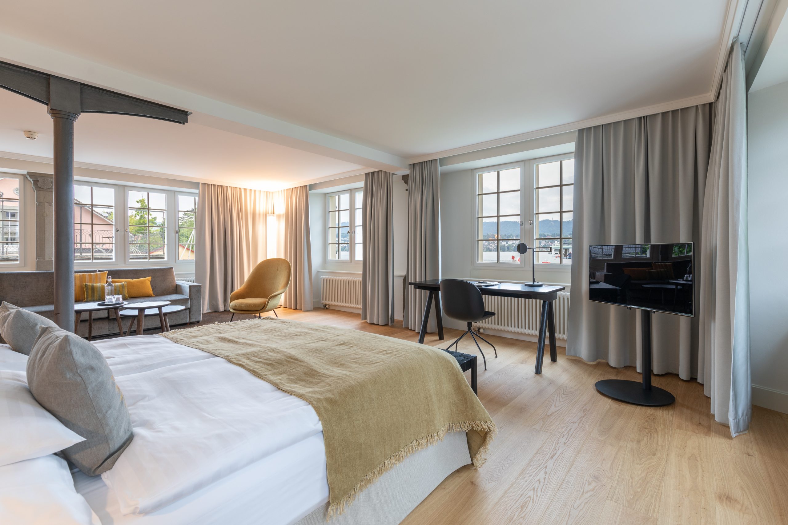 Helles, modernes Boutiquehotelzimmer mit großen Fenstern, Doppelbett mit beigefarbener Decke, Sitzecke mit Sofa und gelbem Stuhl, Schreibtisch mit Stuhl, Fernseher auf einem Ständer und hellem Holzboden. Neutrale Töne und natürliches Licht erfüllen den Raum.