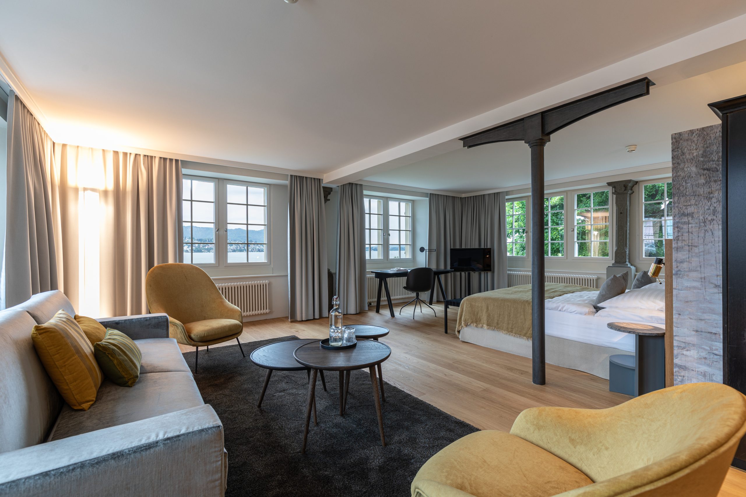 Ein geräumiges, modernes Boutiquehotelzimmer mit großen Fenstern, einem Bett, einem grauen Sofa mit gelben Kissen, einem runden Couchtisch, einem Sessel, einem Schreibtisch und Holzböden. Natürliches Licht füllt den Raum.