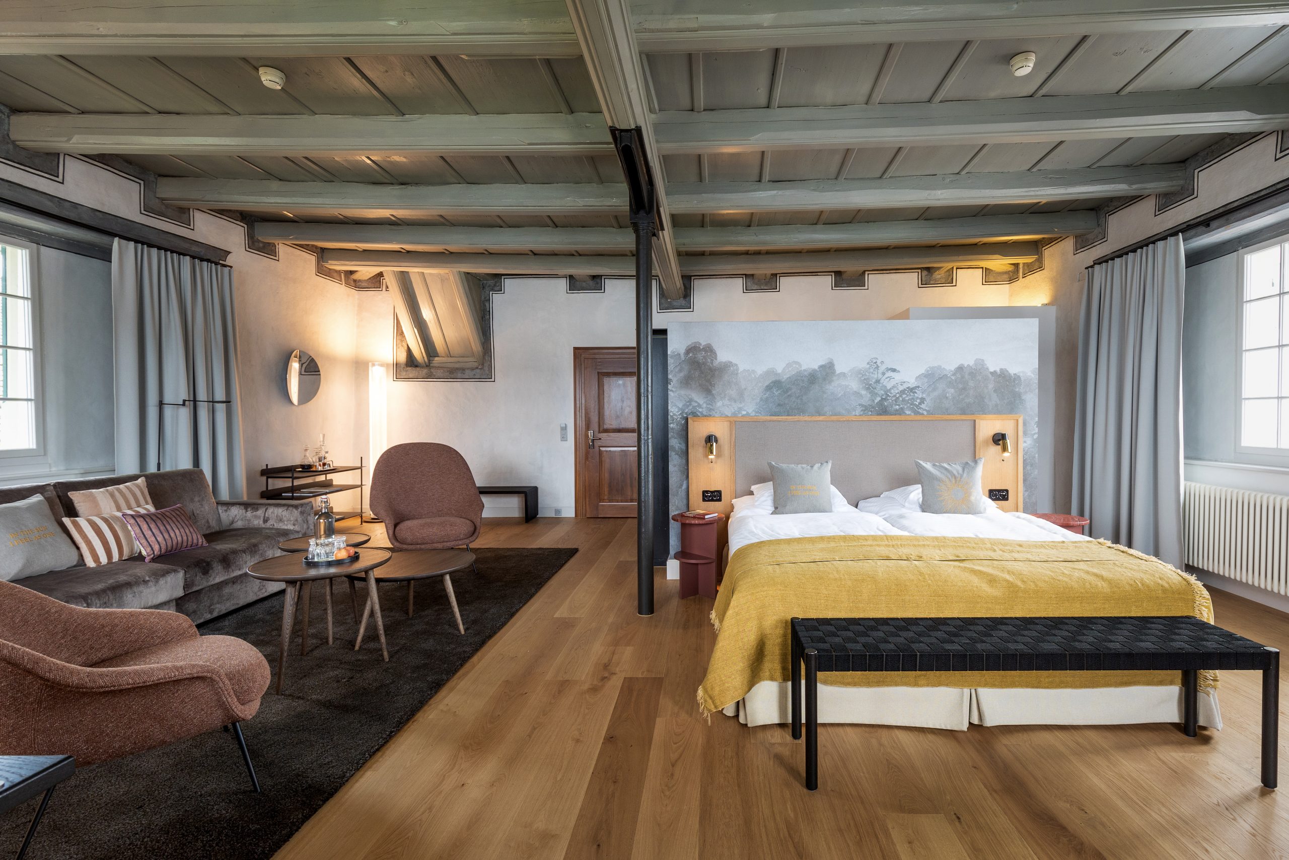 Geräumiges, modernes Boutiquehotel-Zimmer mit sichtbaren Balken, Holzboden, Doppelbett mit gelber Decke, Sitzecke mit Stühlen und Sofa, großen Fenstern und sanfter natürlicher Beleuchtung.