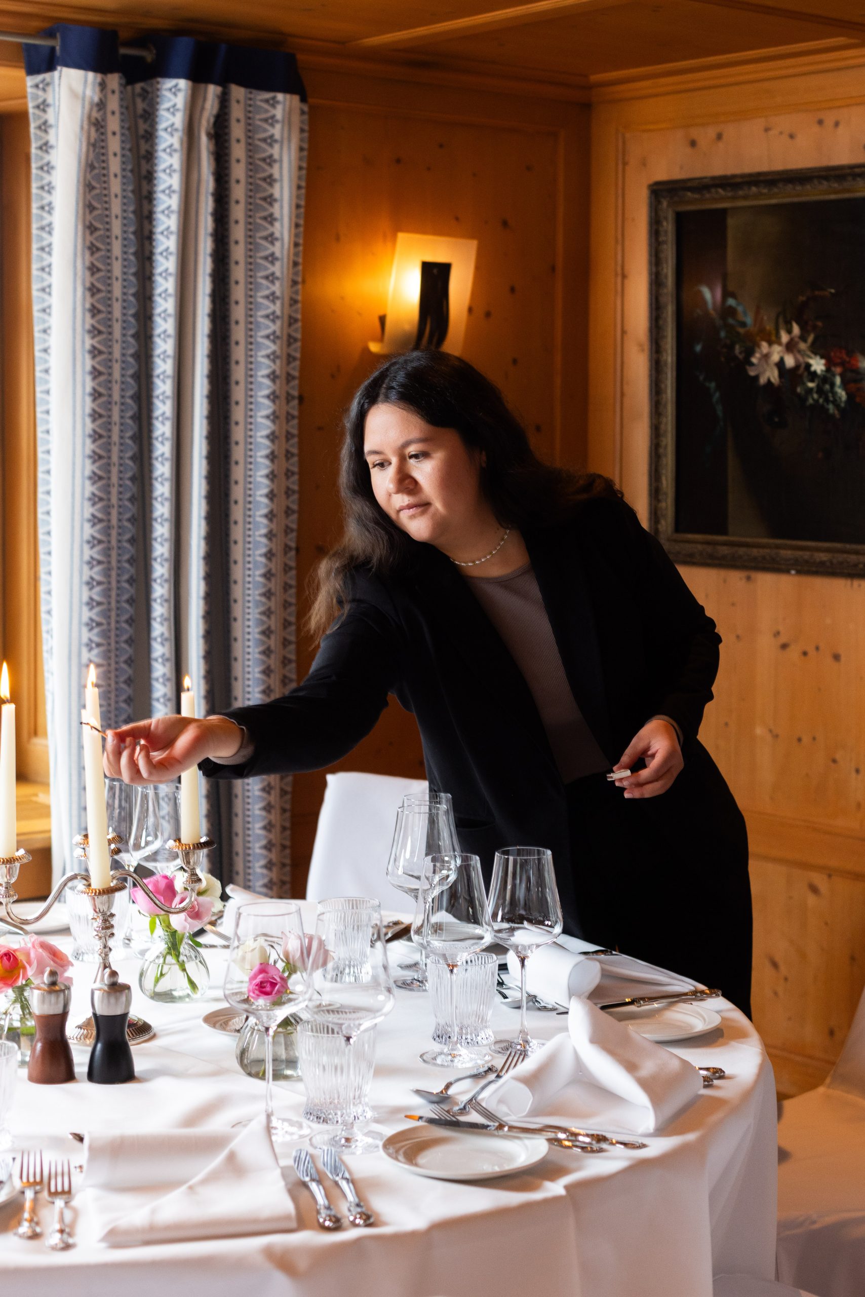 Eine Frau in einem schwarzen Blazer zündet Kerzen auf einem elegant gedeckten Esstisch mit Gläsern, Silberbesteck und Blumendekorationen in einem warm beleuchteten Holzzimmer eines Boutiquehotels an.