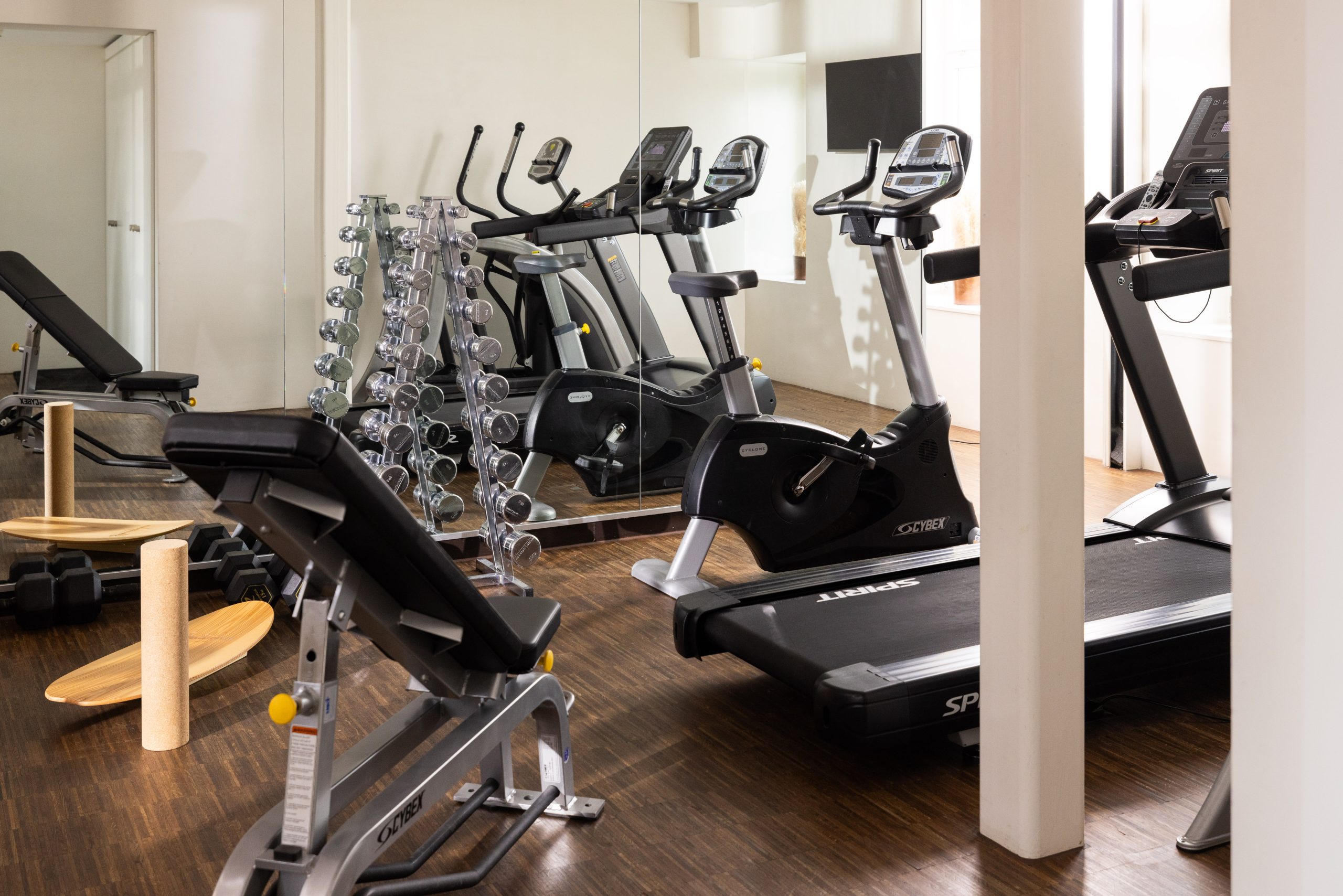 Ein modernes Fitnessstudio in einem Boutique-Hotel mit Laufbändern, stationären Fahrrädern, einer Bank, Hanteln auf einer Ablage, Balance-Boards und Spiegeln, in denen sich die Geräte spiegeln - alles auf einem stilvollen Holzboden.