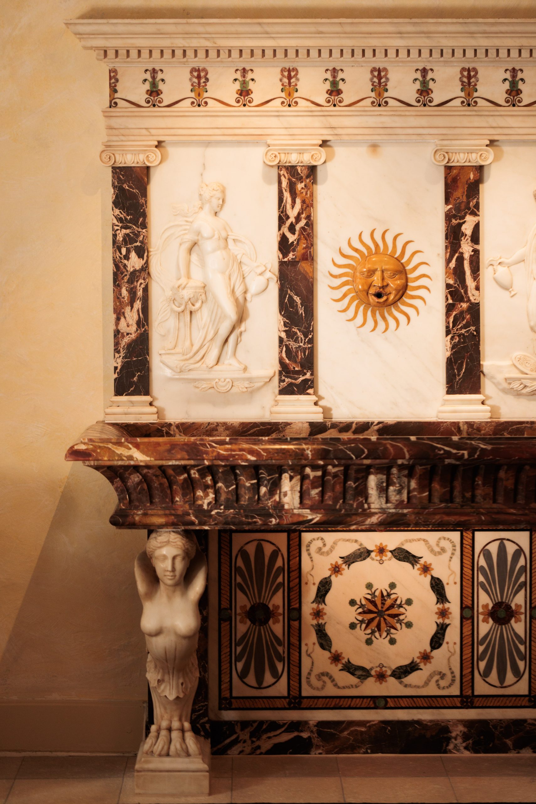 Dekorativer Marmorkamin mit kunstvollen Schnitzereien, darunter eine weibliche Figur, ein Sonnenmotiv, florale Motive und eine Frauenskulptur, die den Kaminsims stützt - perfekt, um die Eleganz eines Boutiquehotels in weißem, braunem und goldenem Steinwerk zu unterstreichen.