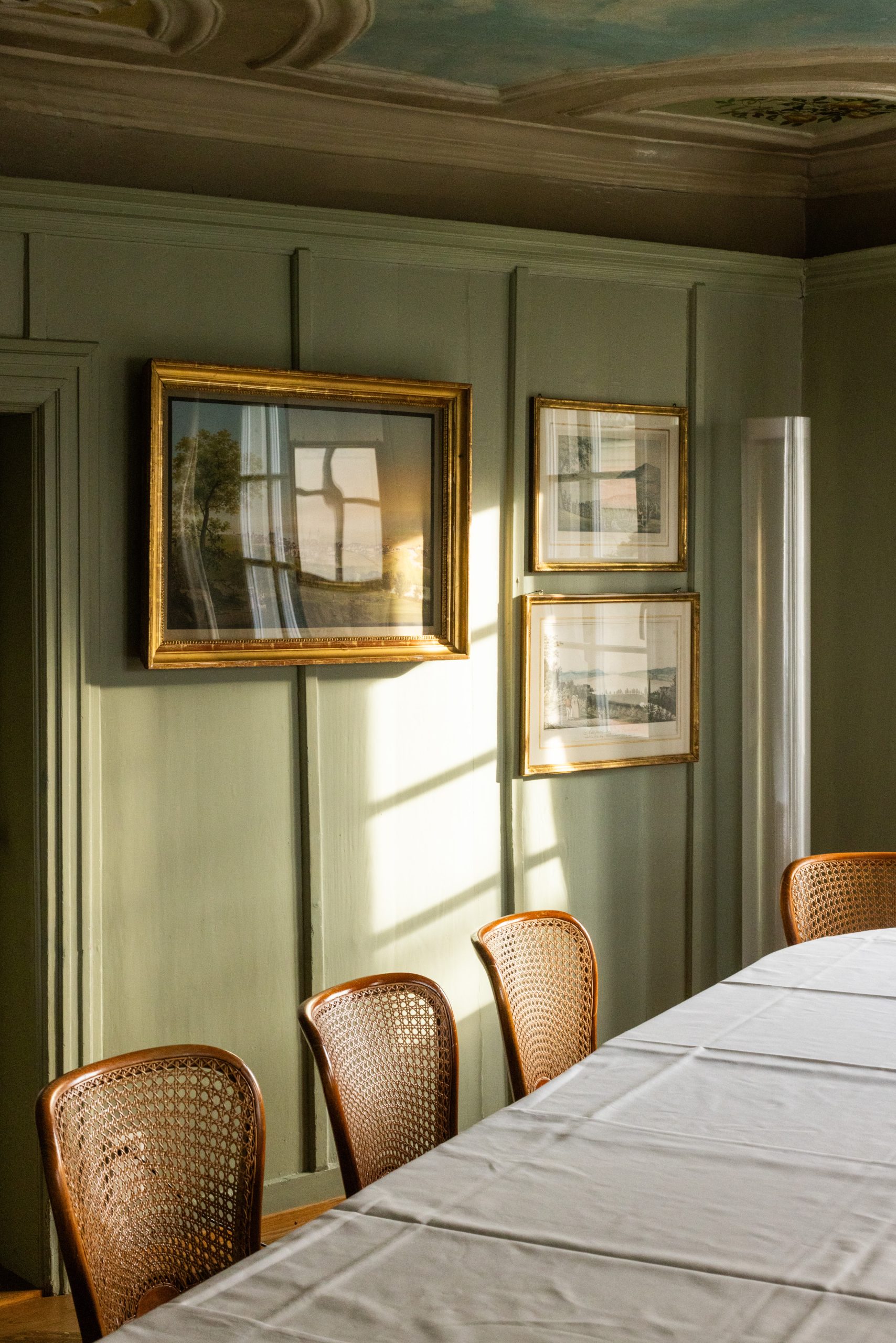 Ein sonnendurchfluteter Speisesaal in einem Boutiquehotel mit Stühlen mit Rohrrücken, weißem Tischtuch und gerahmten Kunstwerken an hellgrün getäfelten Wänden. Das Sonnenlicht strömt durch das Fenster und wirft sanfte Schatten an die Wand.