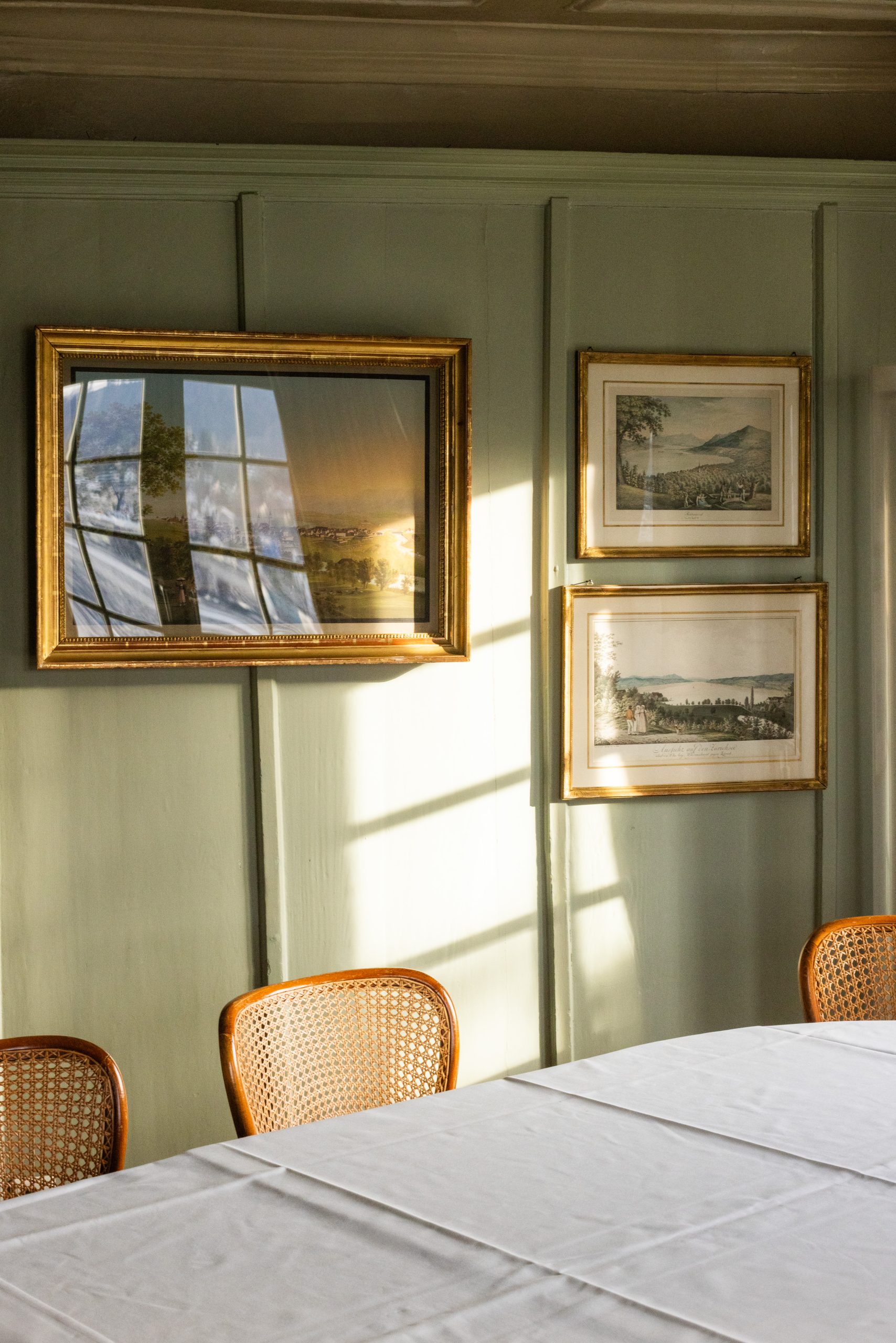 Ein sanftes, sonnendurchflutetes Zimmer in einem Boutiquehotel mit einer mintgrünen Wand, an der drei gerahmte Landschaftsgemälde hängen. Im Vordergrund sieht man einen mit einem weißen Tuch bedeckten Tisch und drei Stühle mit Korbrücken.