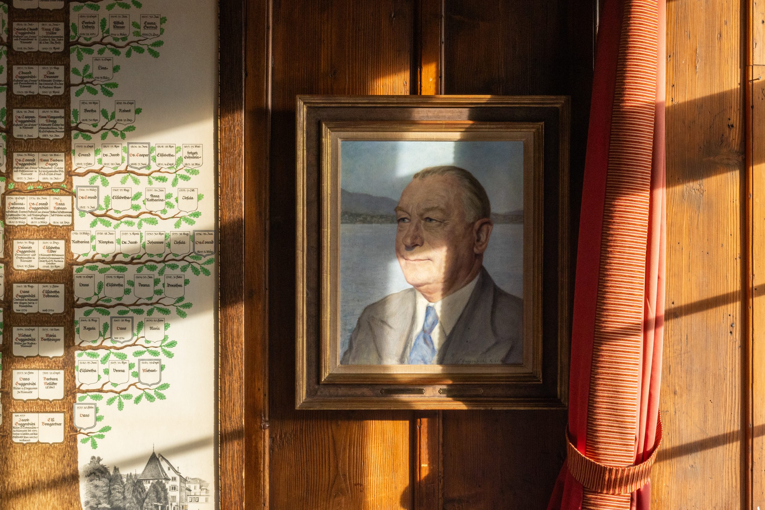Das gerahmte Porträt eines älteren Mannes im Anzug hängt an einer Holzwand, die vom Sonnenlicht in diesem Boutiquehotel beleuchtet wird. Links ist ein Familienstammbaum abgebildet, und rechts verdeckt ein roter Vorhang teilweise das Fenster.